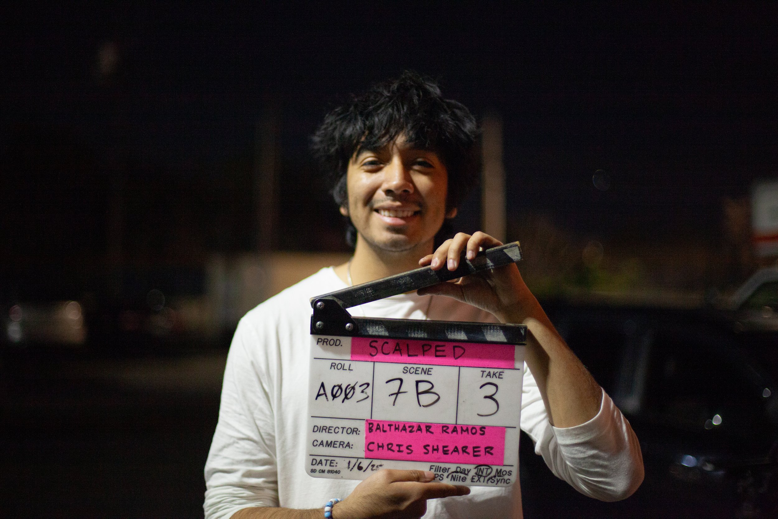 A young man with curly black hair smiling, holding a clapperboard with pink labels on it, in an outdoor nighttime setting.