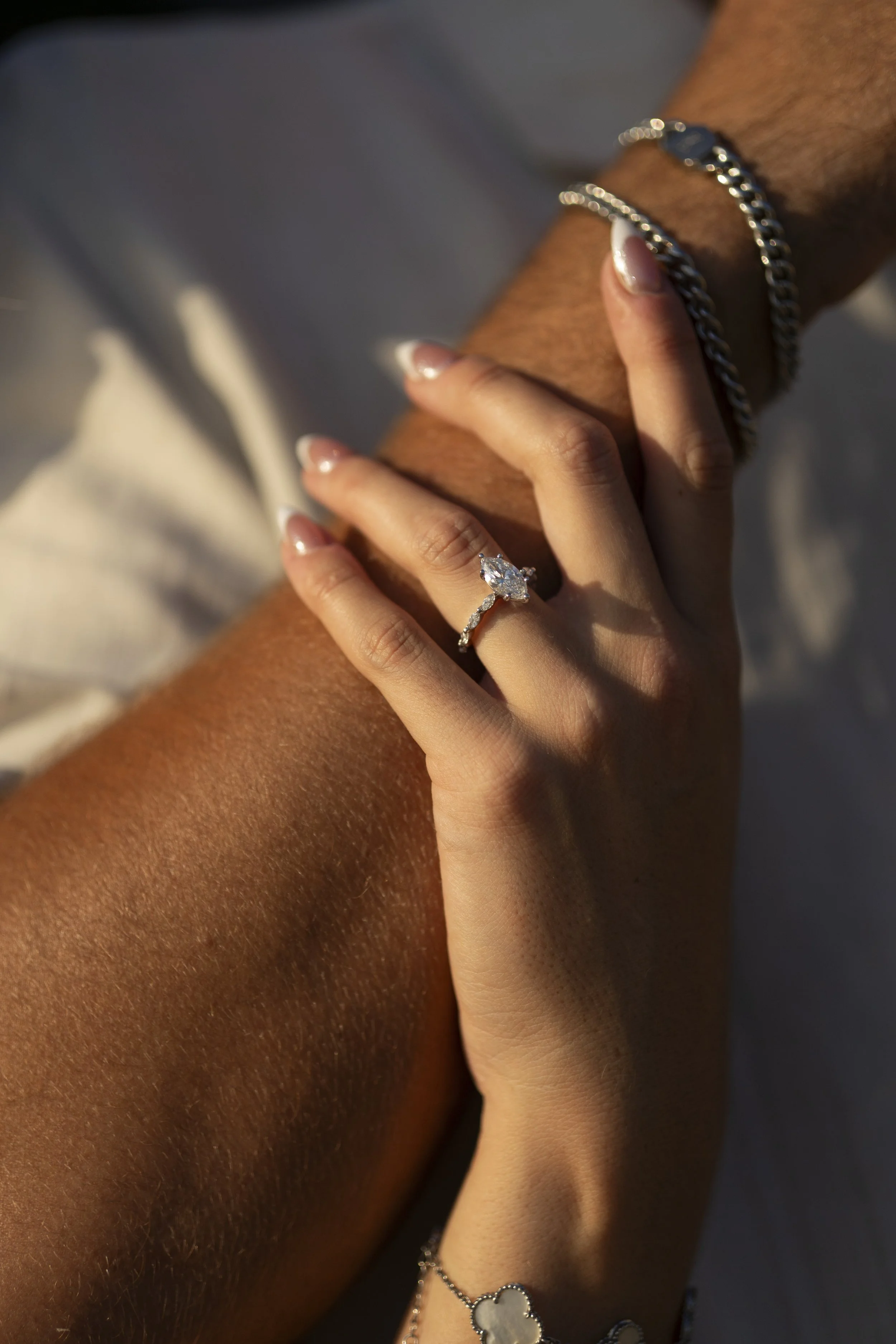 Close-up of two hands, one of darker skin and the other lighter, intertwined. The person with lighter skin is wearing an engagement ring with a large central diamond and delicate band. The darker-skinned person is wearing silver jewelry, including a chain bracelet and a flower-shaped ring. The background is out of focus.