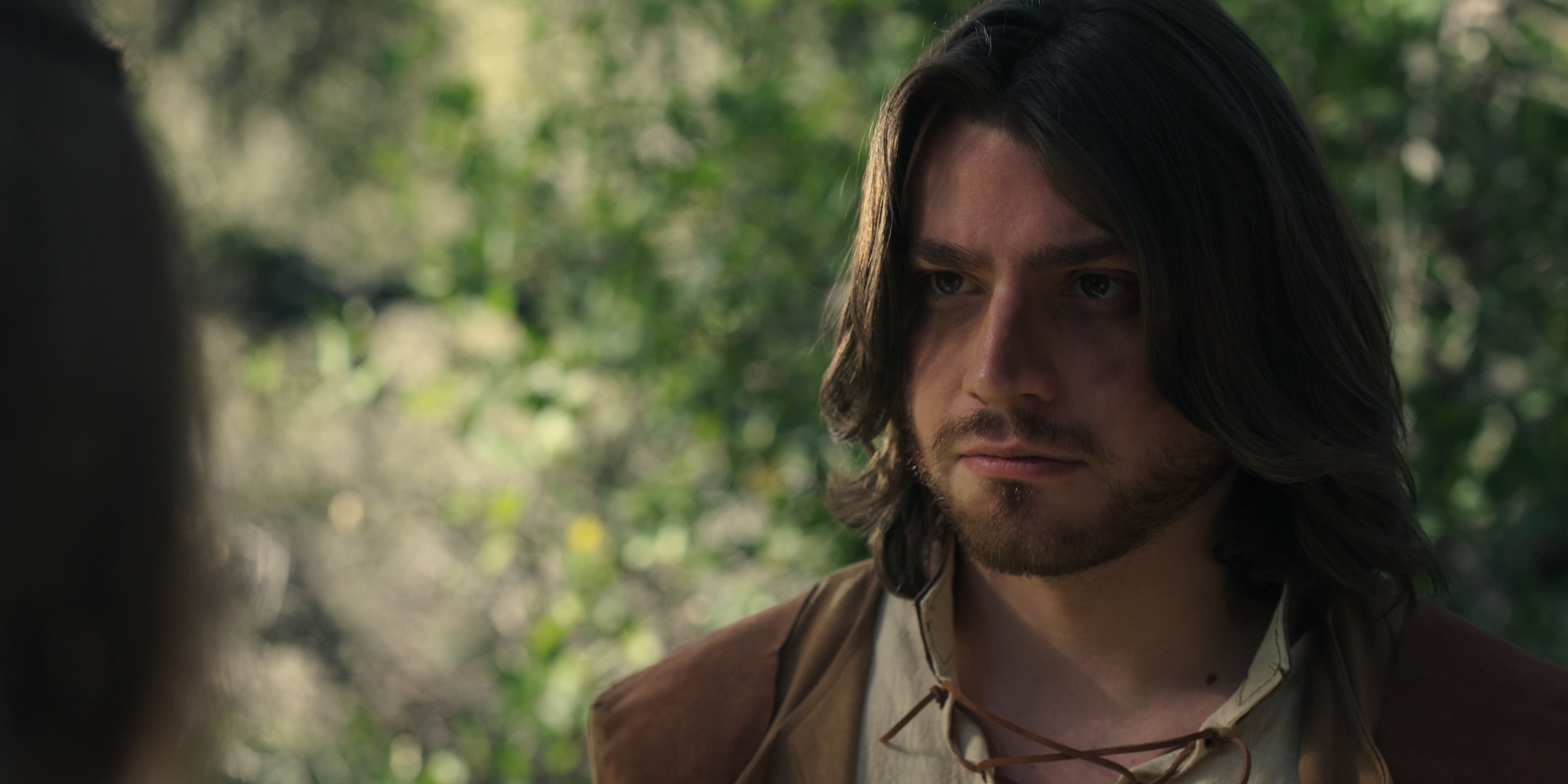 Close-up of a young man with long dark hair and a beard, wearing brown clothing, standing outdoors with a blurred green leafy background.