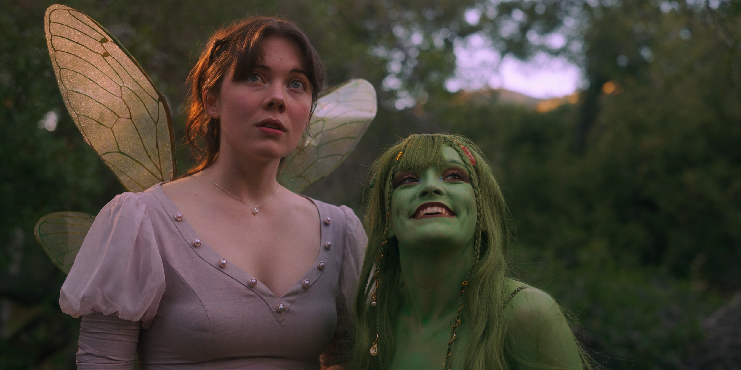 Two women dressed as fairies, one with butterfly wings and the other with green face paint and long hair, standing outdoors in a wooded area.