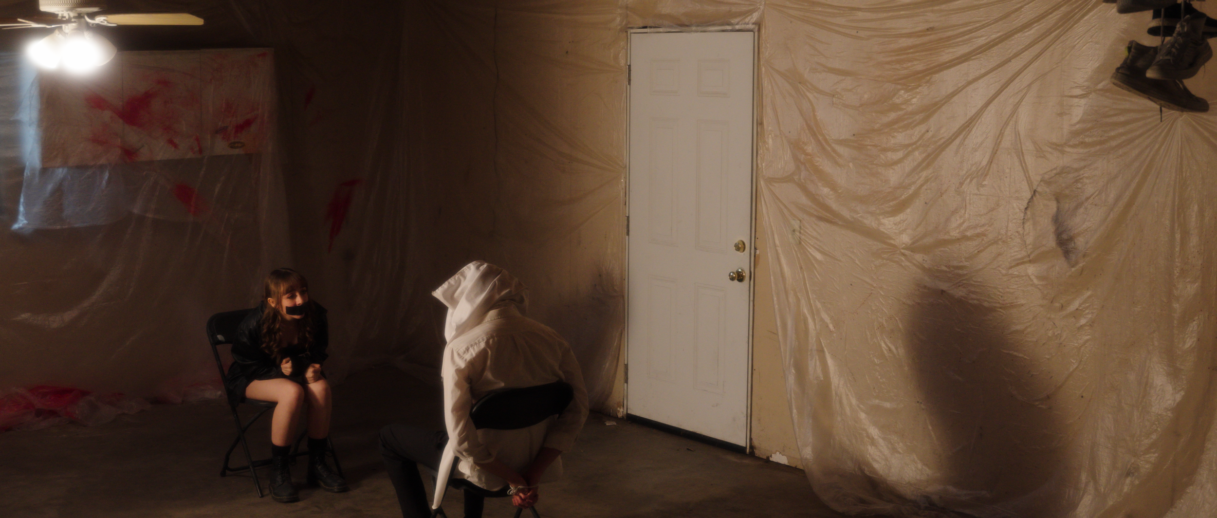 A girl with a black face mask sitting on a chair, a person in a white hoodie crouching, and another person in white hoodie with horse mask in a room with plastic-covered walls and a closed door.