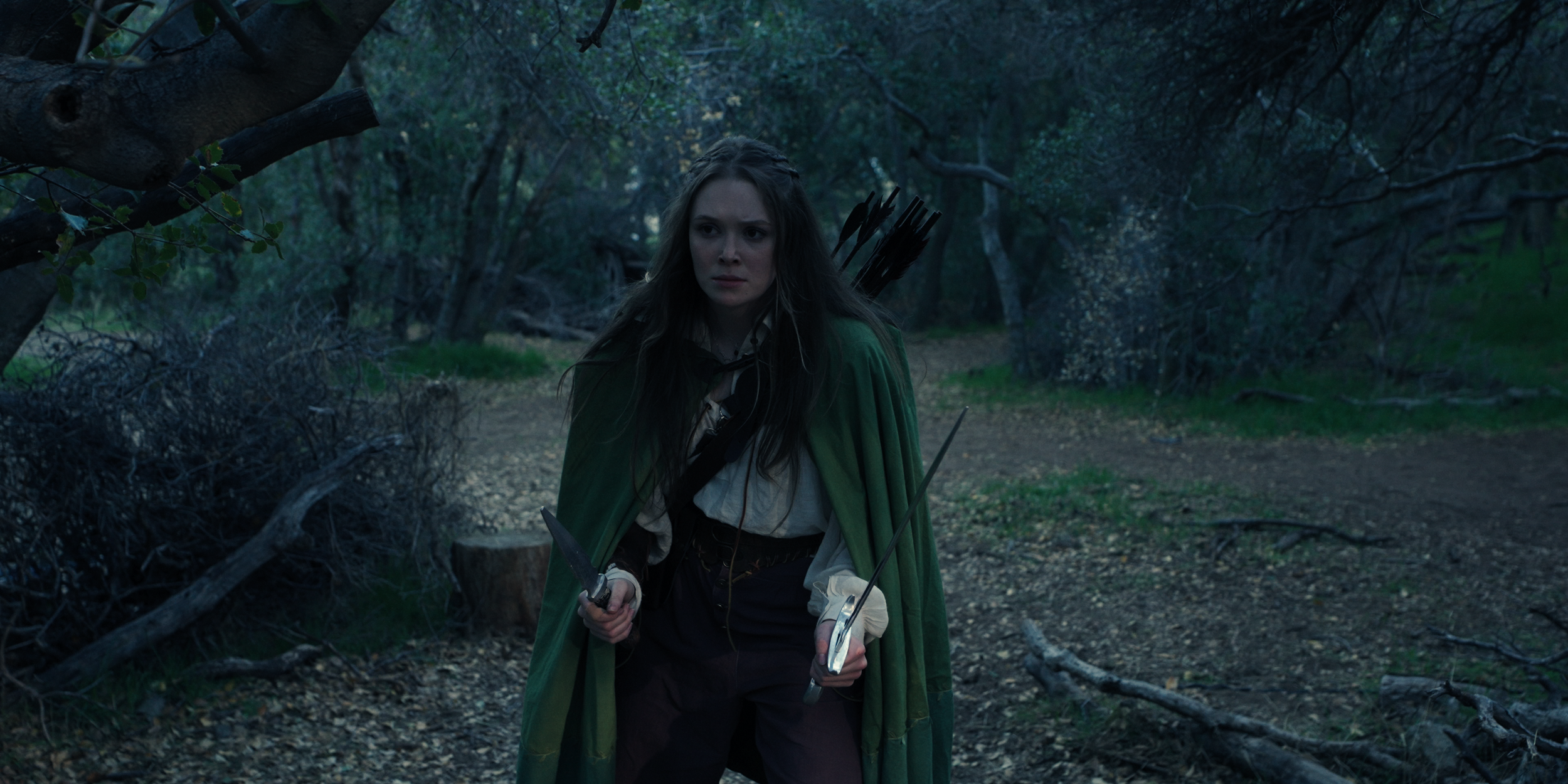 A woman dressed as an archer with a cape and arrows on her back walks through a dark, shadowy forest holding a bow and arrow.