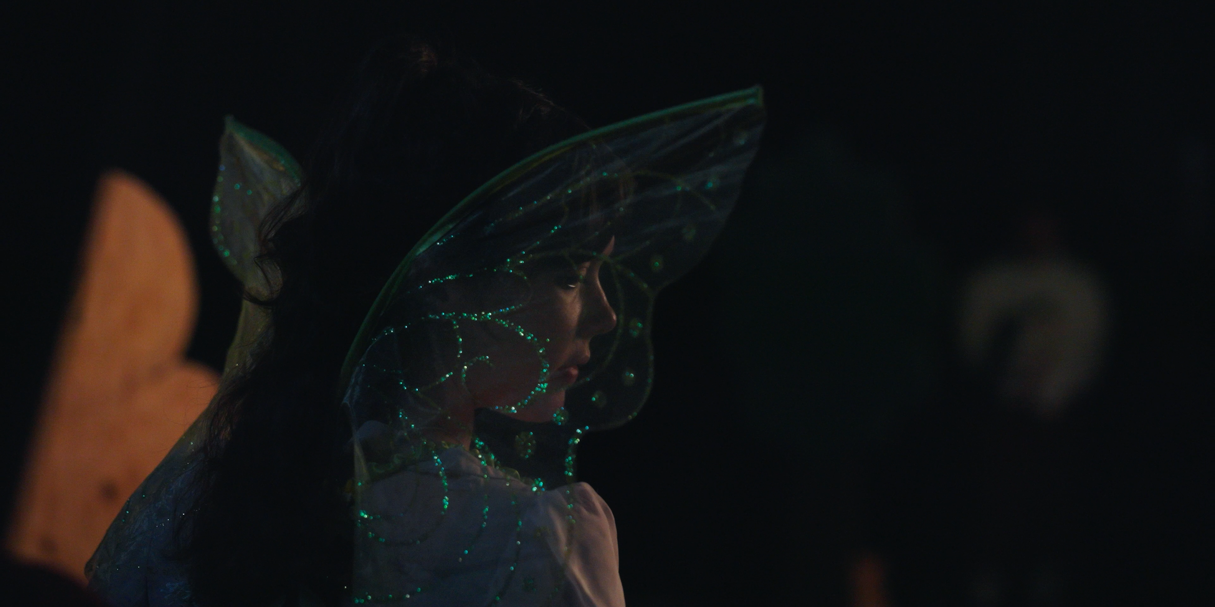 A woman wearing glowing, iridescent fairy wings, illuminated faintly in the dark.