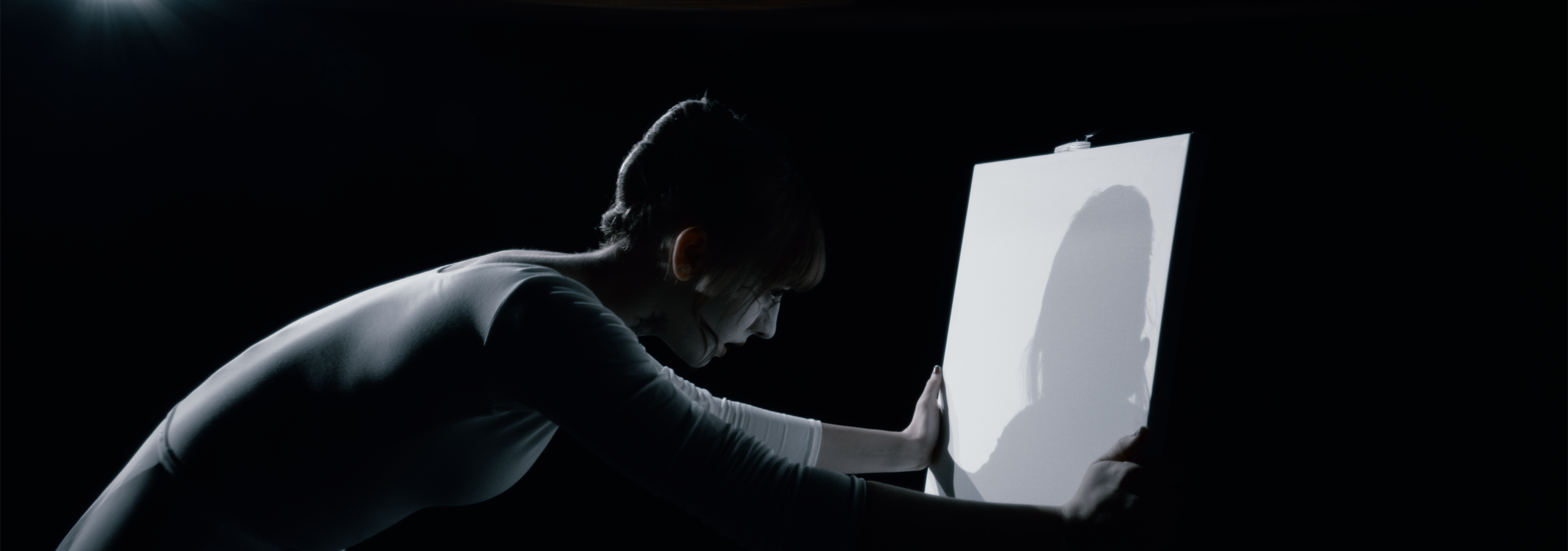 A person in a dark room is pressing their hands against a bright, illuminated canvas, creating a shadow on it.