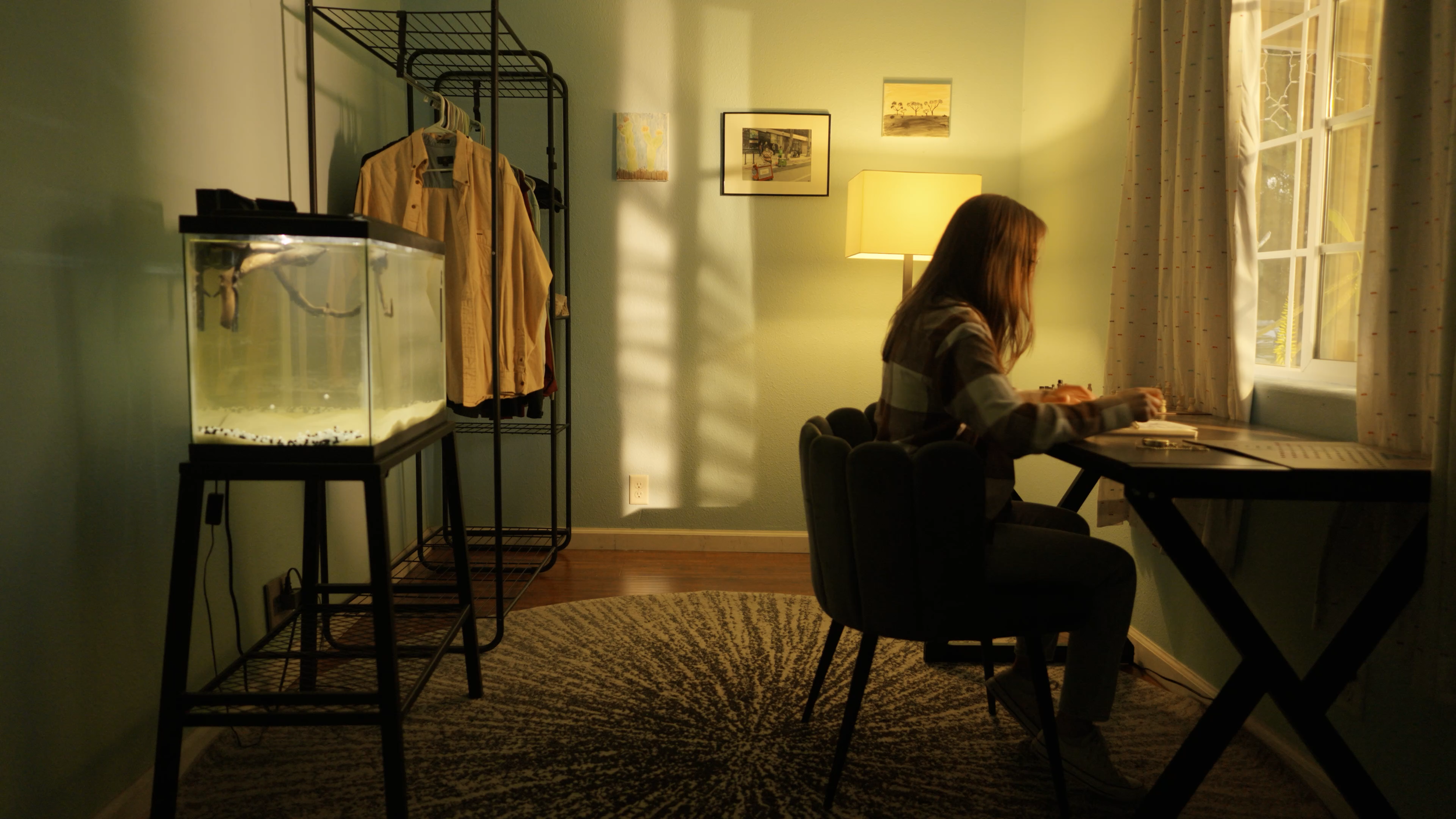 A woman with long hair sitting at a desk by a window, working or studying in a cozy room illuminated by a yellow lamp, with a fish tank and clothing rack nearby.