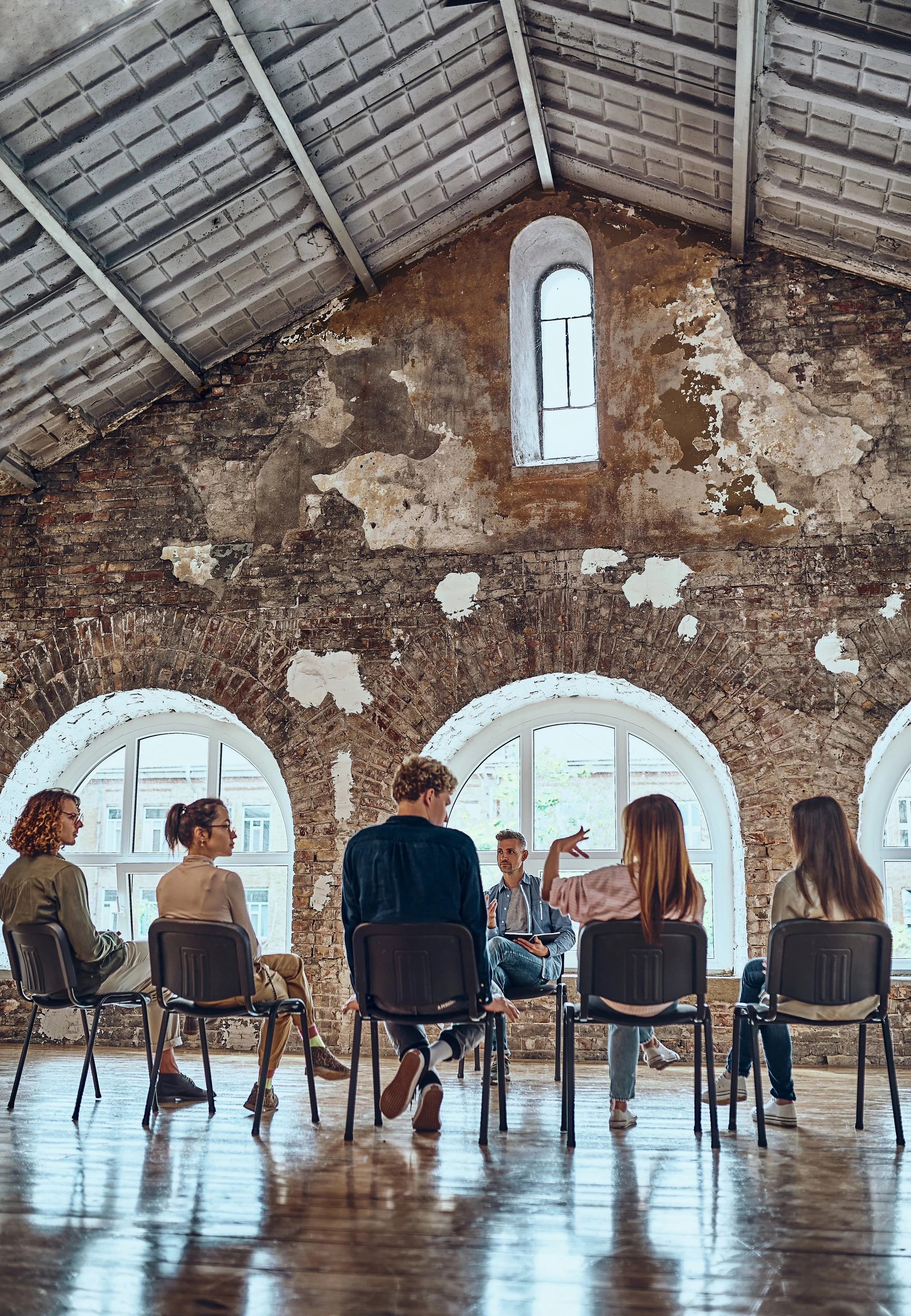A group of people in a meeting or discussion inside a large, rustic industrial-style room with brick walls and arched windows. The participants are sitting on chairs forming a circle, with some actively engaging in the conversation.