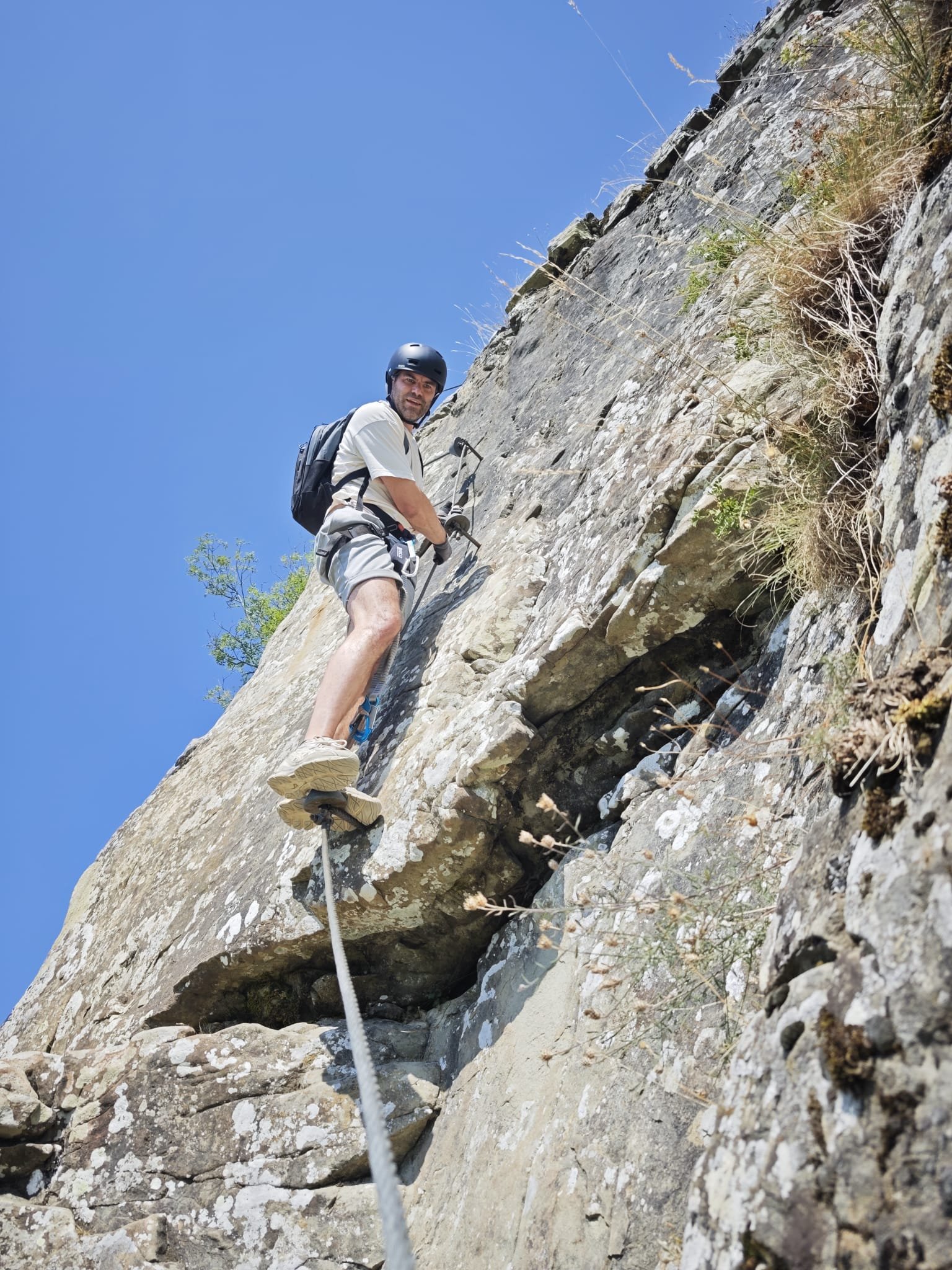 A man wearing a helmet and hiking gear is rock climbing on a steep, rocky cliff using safety ropes under a clear blue sky.