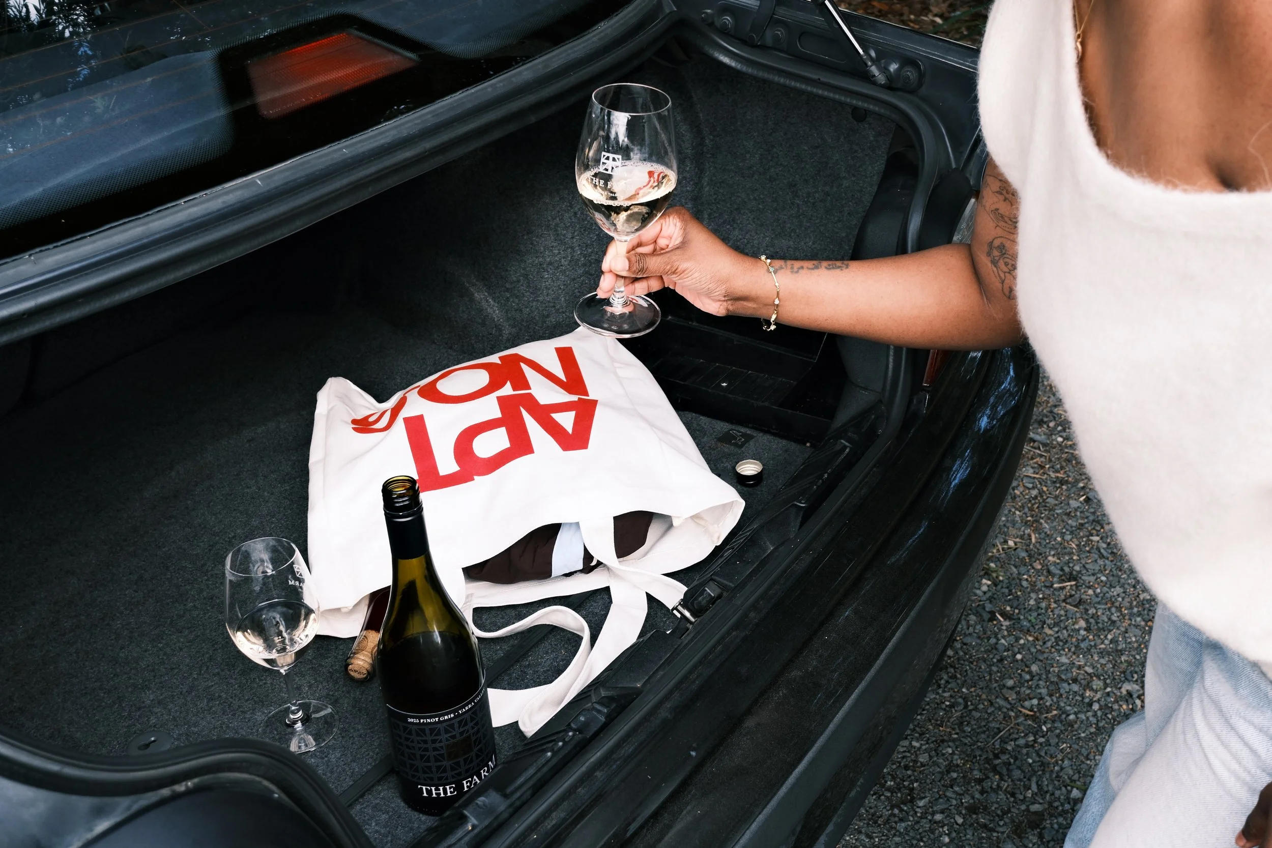 Person holding a glass of white wine or champagne in the trunk of a car with empty wine bottles, a white tote bag with red text, and a wine glass on the ground nearby.