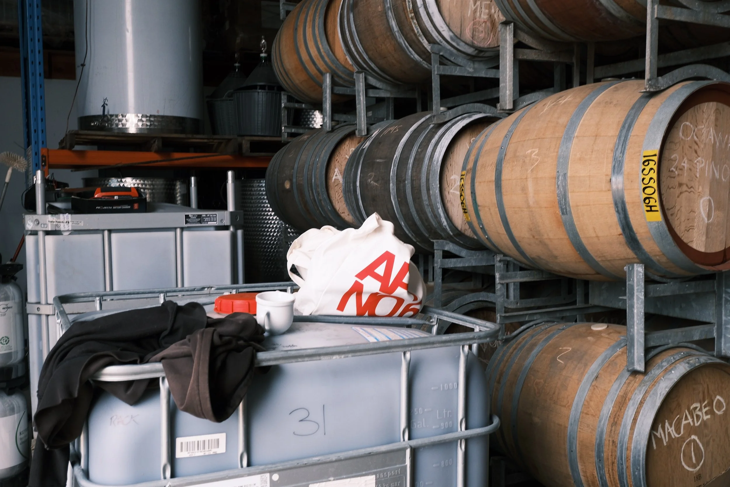 Wooden barrels with metal bands stored on metal racks in a warehouse or storage room. Several items such as a plastic storage container, a bag, and miscellaneous objects are placed on a gray metal cart in the foreground.