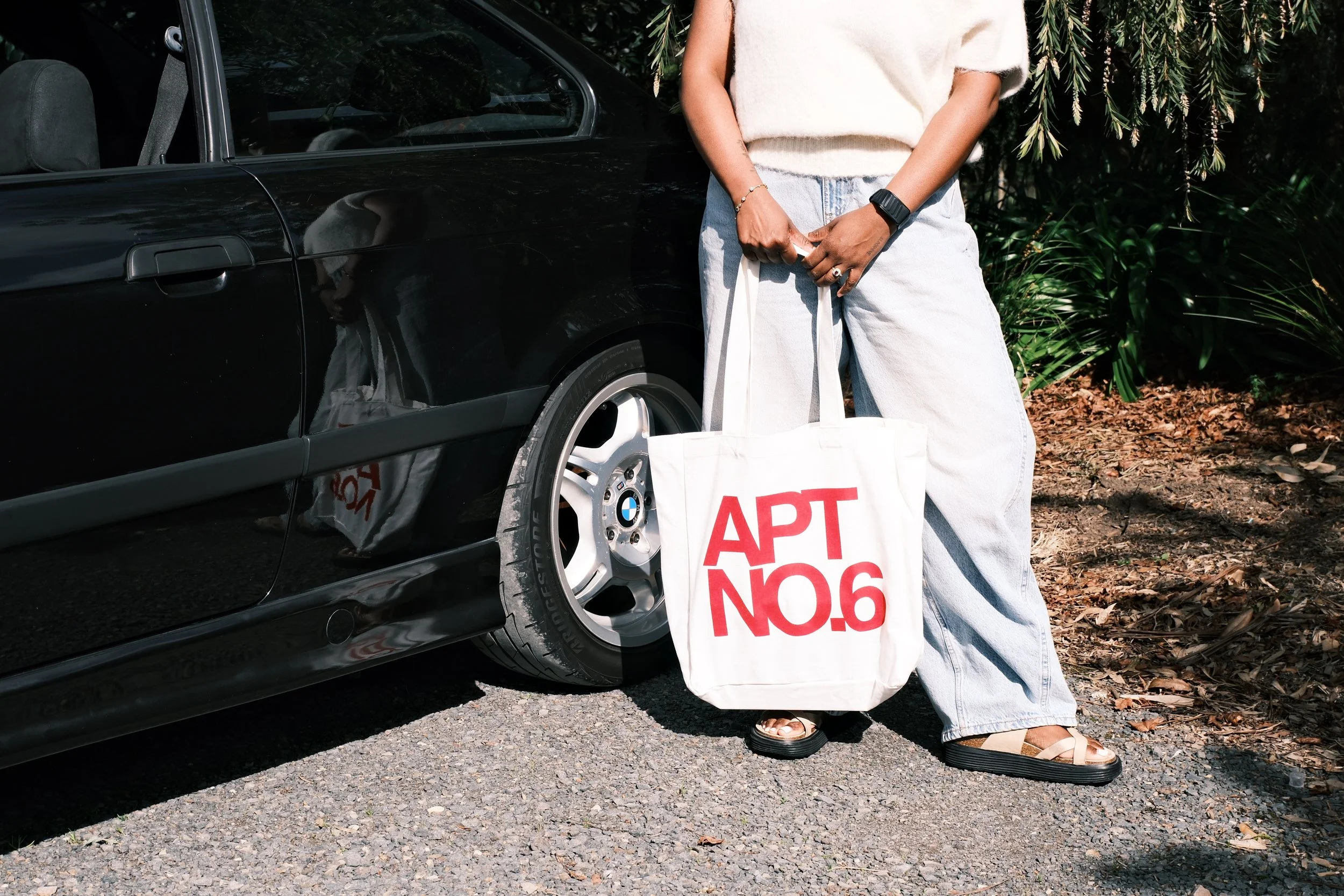A person standing next to a black car holding a white tote bag with red text. The person's face is not visible, wearing a white top, light blue jeans, sandals, and a watch. There are trees and foliage in the background.