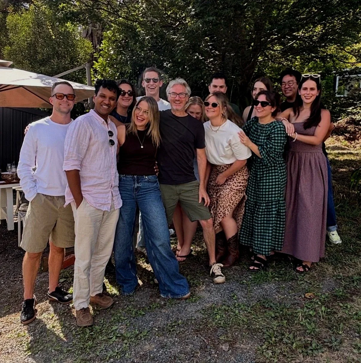 Group of friends smiling and posing outdoors on a sunny day, standing on grass with trees and a patio umbrella in the background.