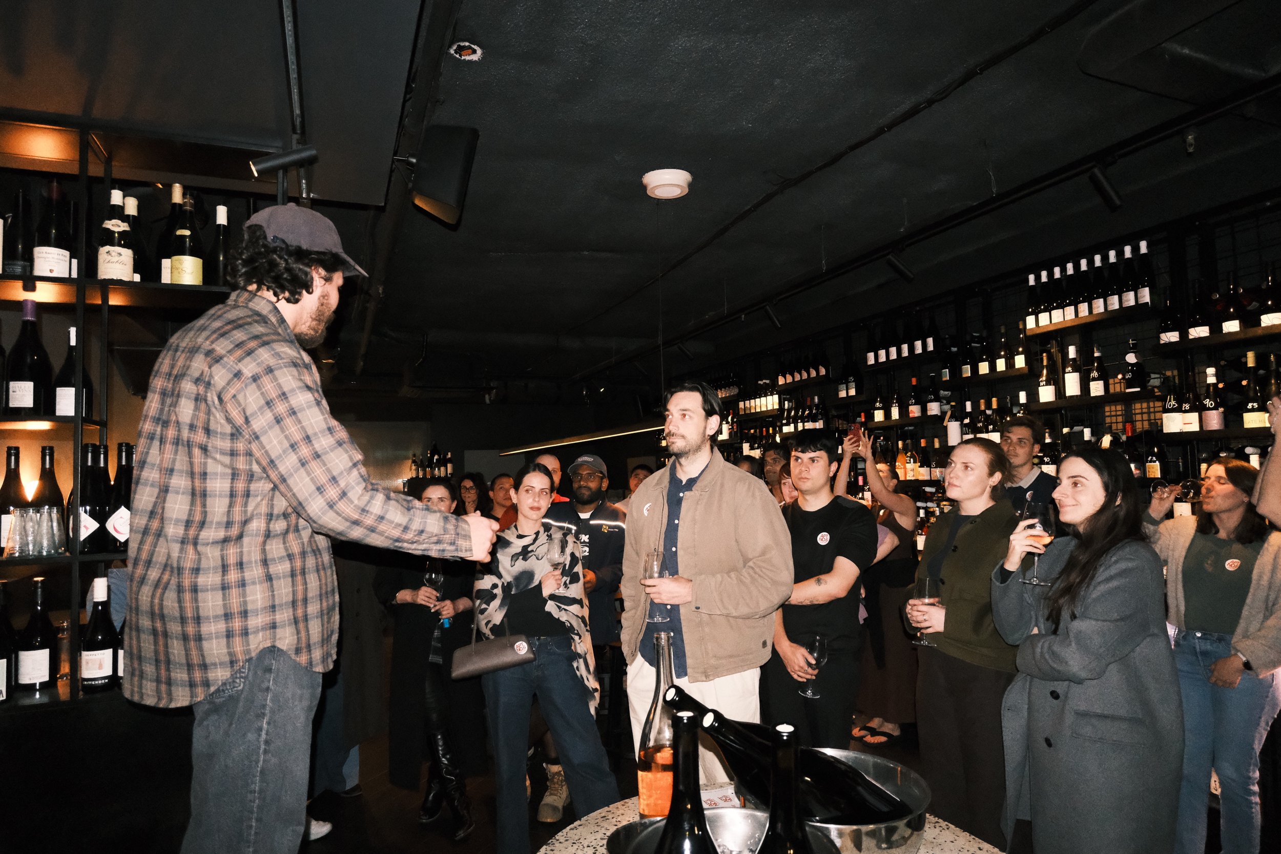 A bartender is serving drinks to a group of people in a bar or wine cellar with shelves of wine bottles on the wall. The crowd is watching, some holding glasses of wine or beer, and the atmosphere appears lively.