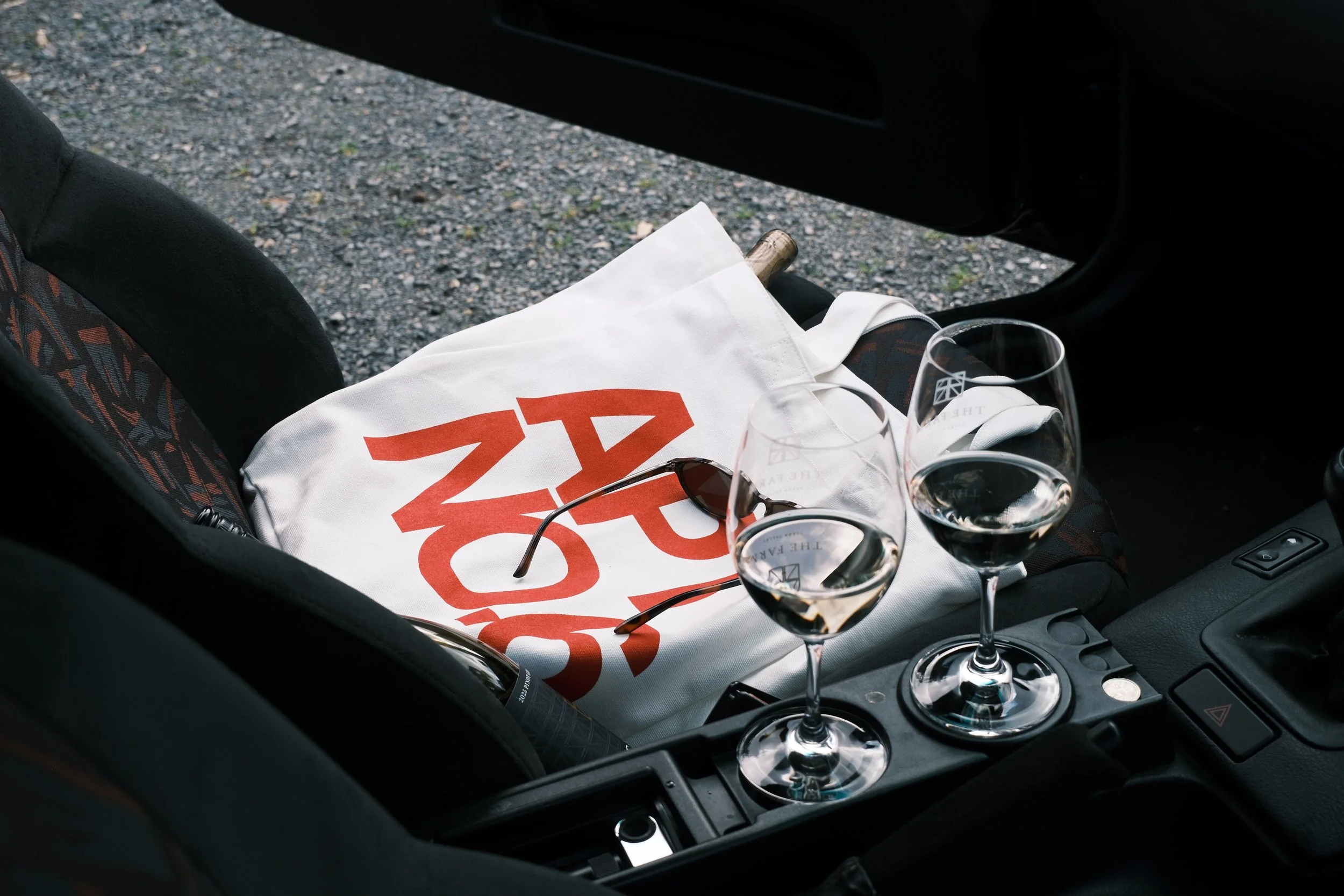 Car interior with a car seat, a white picnic bag with red text, sunglasses, three empty wine glasses, and some coins on the center console. Outside the car, gravel ground is visible.