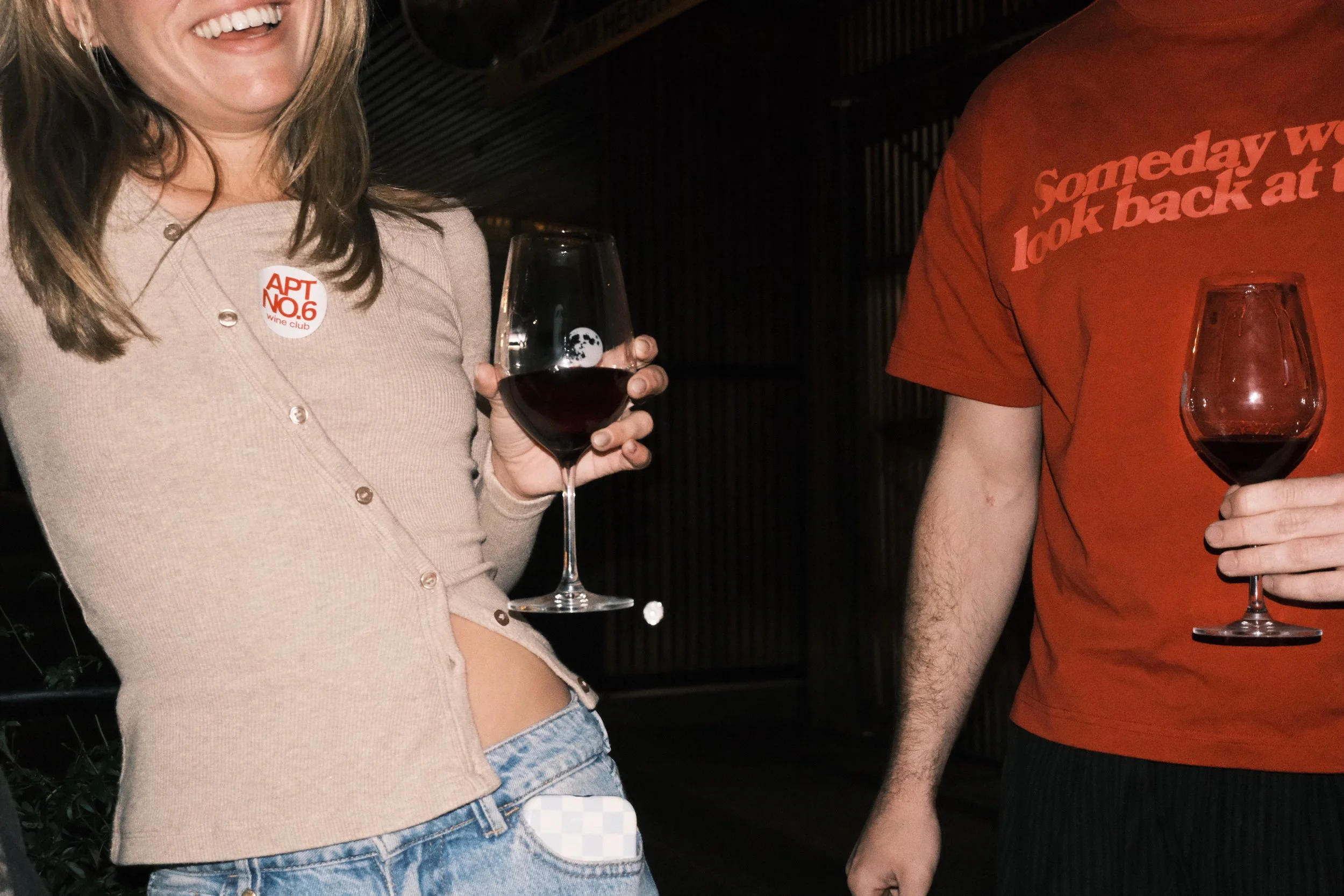 A woman with brown hair holding a glass of red wine, smiling, wearing a beige cardigan with a button that says 'APT NO. 6 wine club'. Next to her, a man in an orange T-shirt with red text holding a glass of red wine, their faces are not visible.