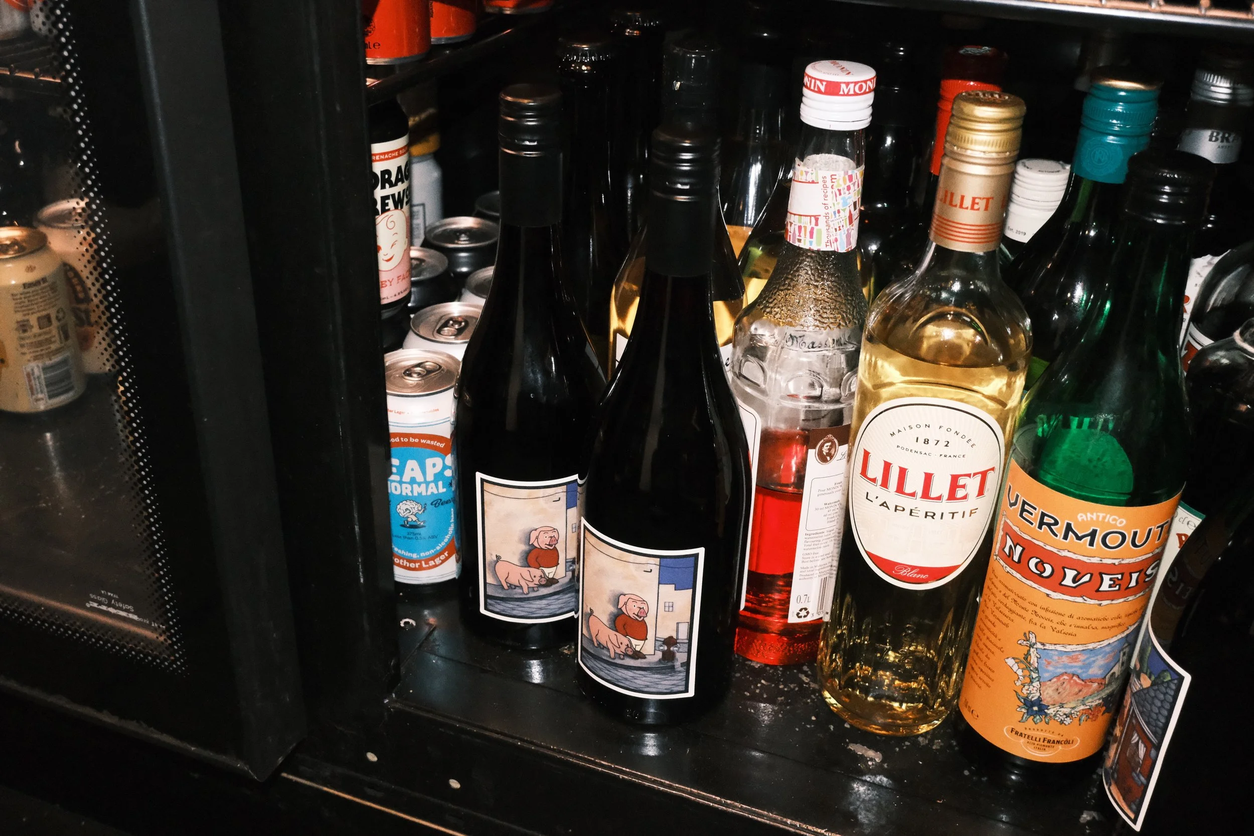 A variety of liquor bottles and cans stored on a shelf inside a refrigerator. The bottles include wine, spirits, and flavored liqueurs, with some labels visible.