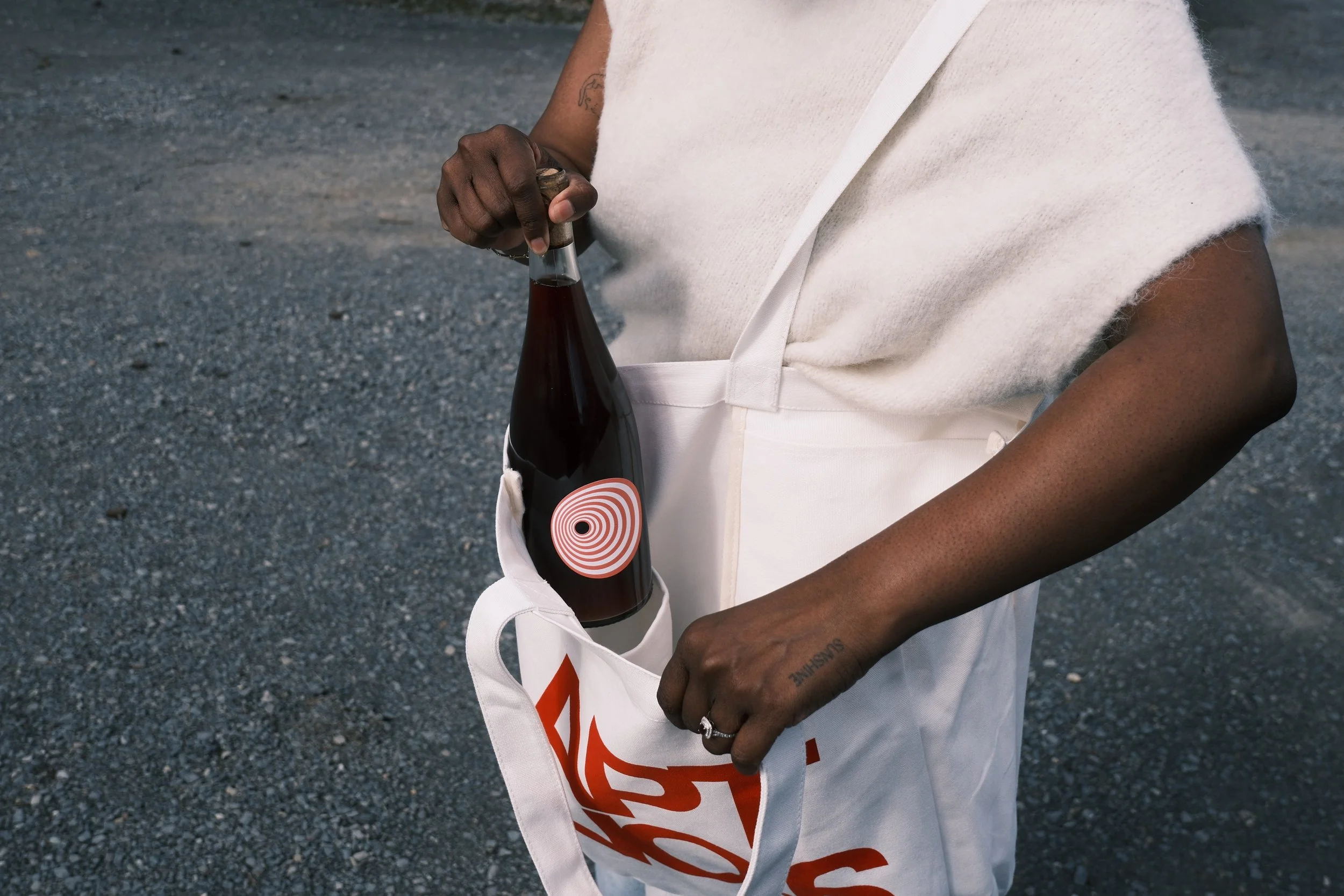 Person holding a bottle of soda and a white tote bag with a red spiral design, standing on a gravel surface.