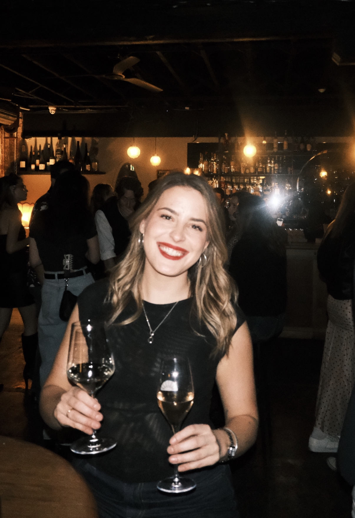 A woman with blonde wavy hair wearing a black dress, smiling with red lipstick, holding two glasses of white wine at a crowded bar or party.