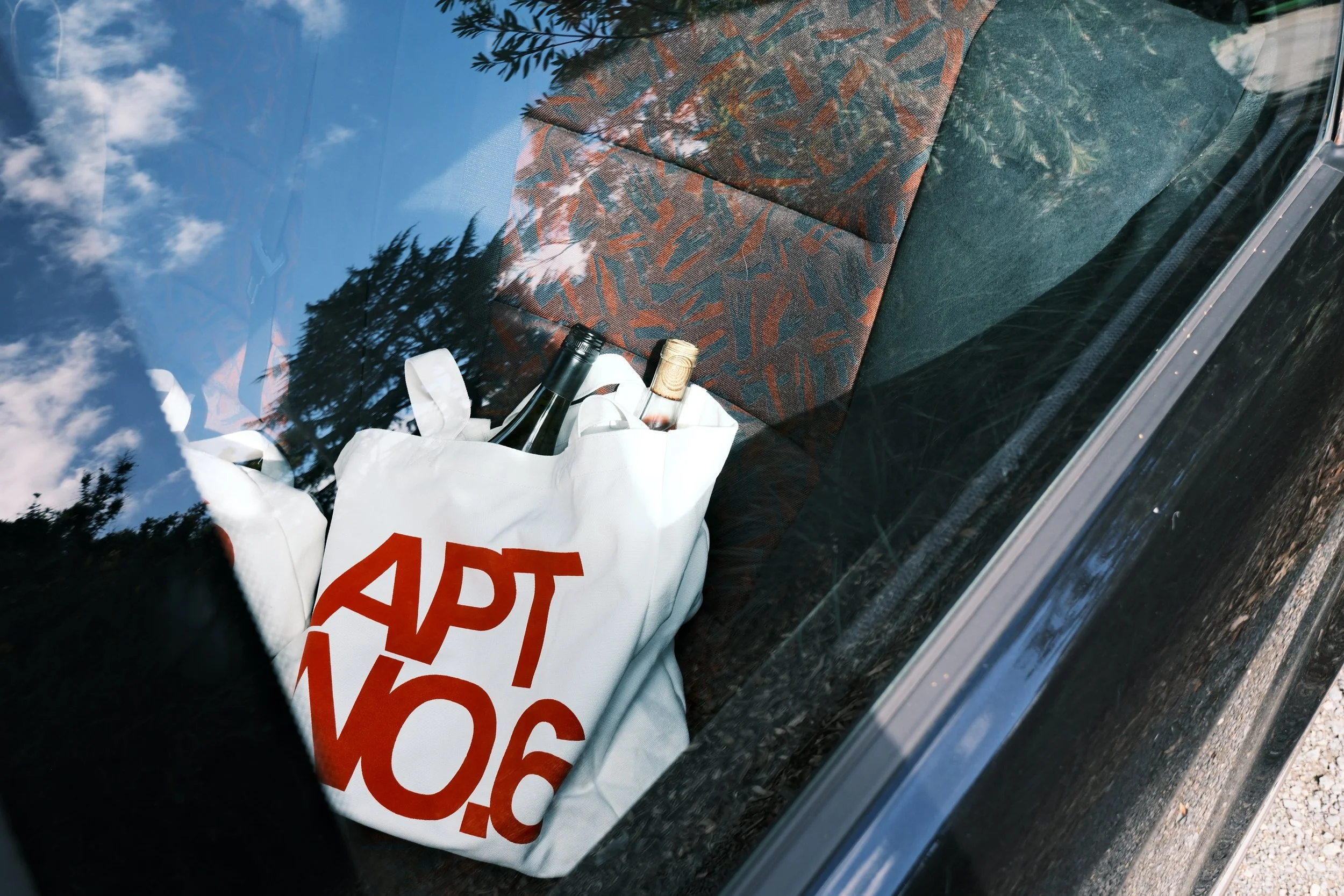 A white paper bag with red text containing bottles, one dark-colored wine bottle with a black cap and another clear bottle with a gold-colored cap, placed on the dashboard of a car. The reflection of blue sky and trees is visible on the car window.