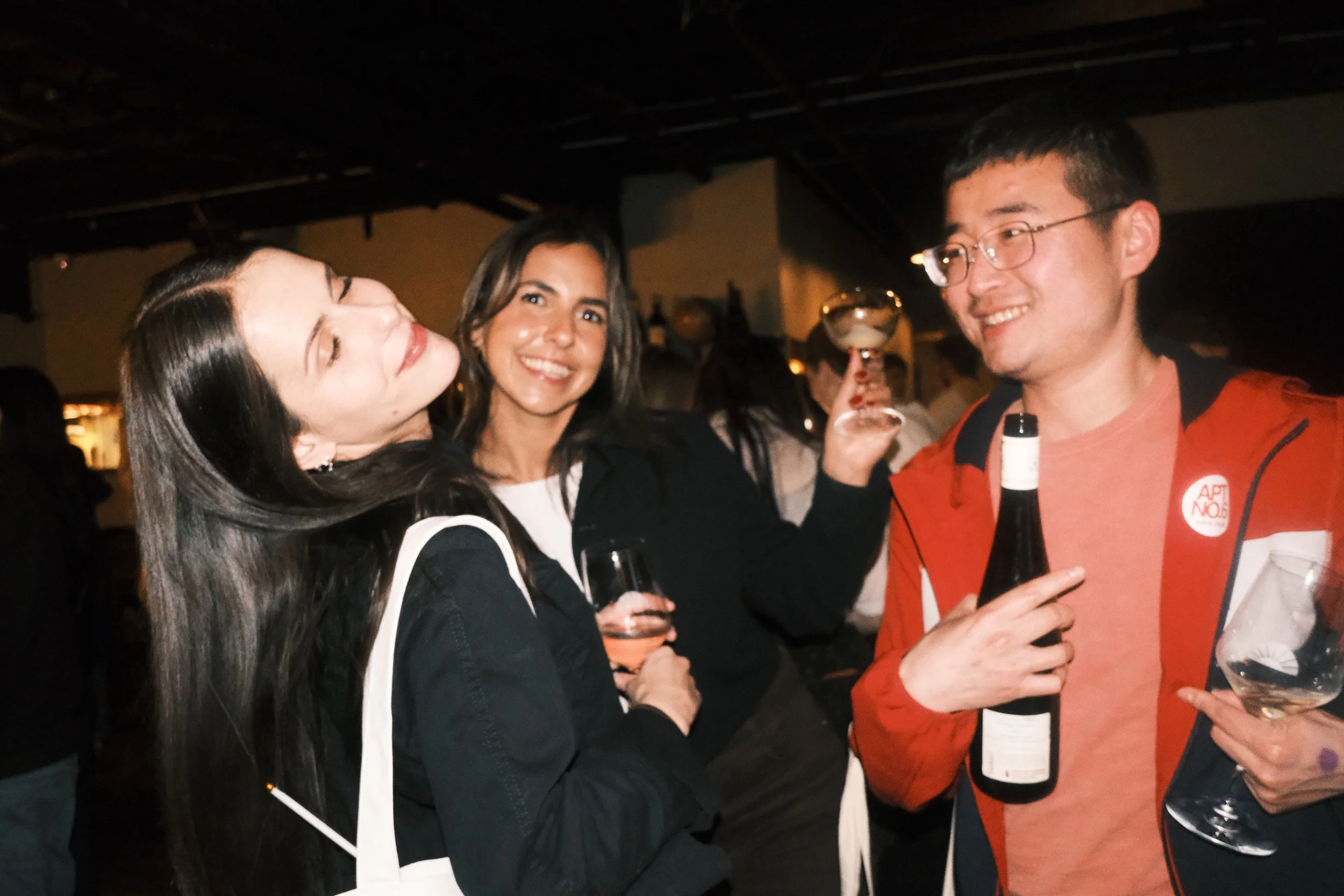 Group of people socializing at a party or gathering, holding drinks, smiling, and enjoying themselves in a dimly lit environment.
