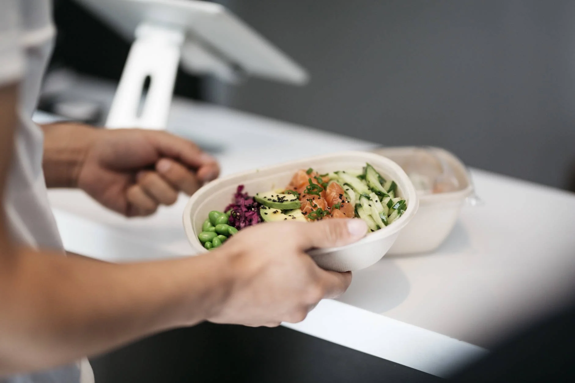 Person holding a bowl of fresh mixed salad with sliced cucumbers, salmon, peas, and black sesame seeds, with food containers in the background.