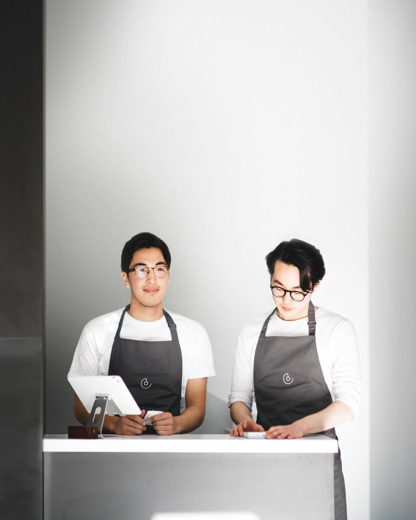 Two young men wearing white shirts, dark aprons, and glasses, working together at a white counter in a minimalist kitchen or food prep area. One is looking at a tablet held in his hands, while the other is focused on something on the counter.