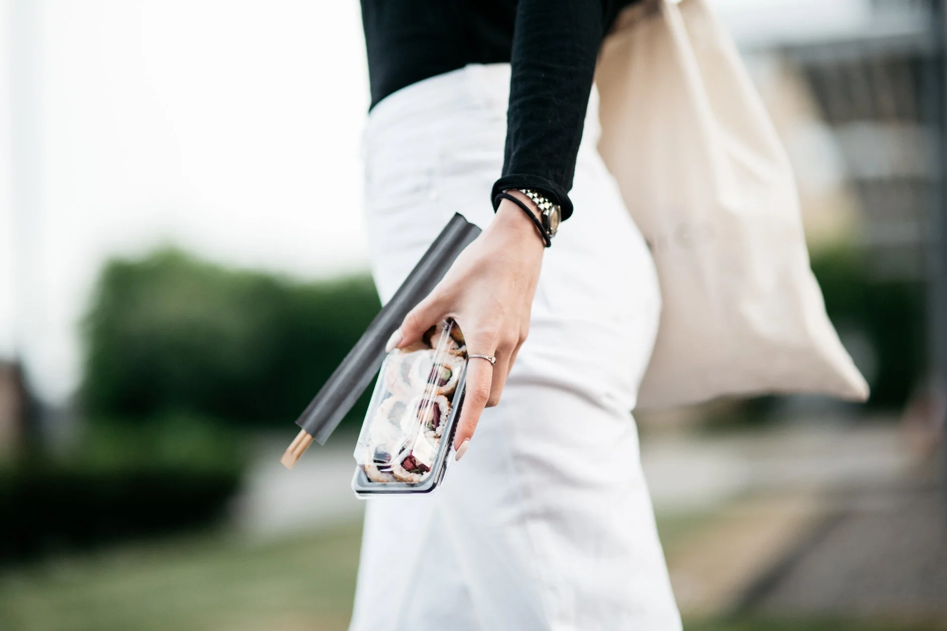 A woman holding chopsticks and a smartphone with a sushi display, wearing white pants, a black top, and a watch, outdoors with blurred greenery in the background.