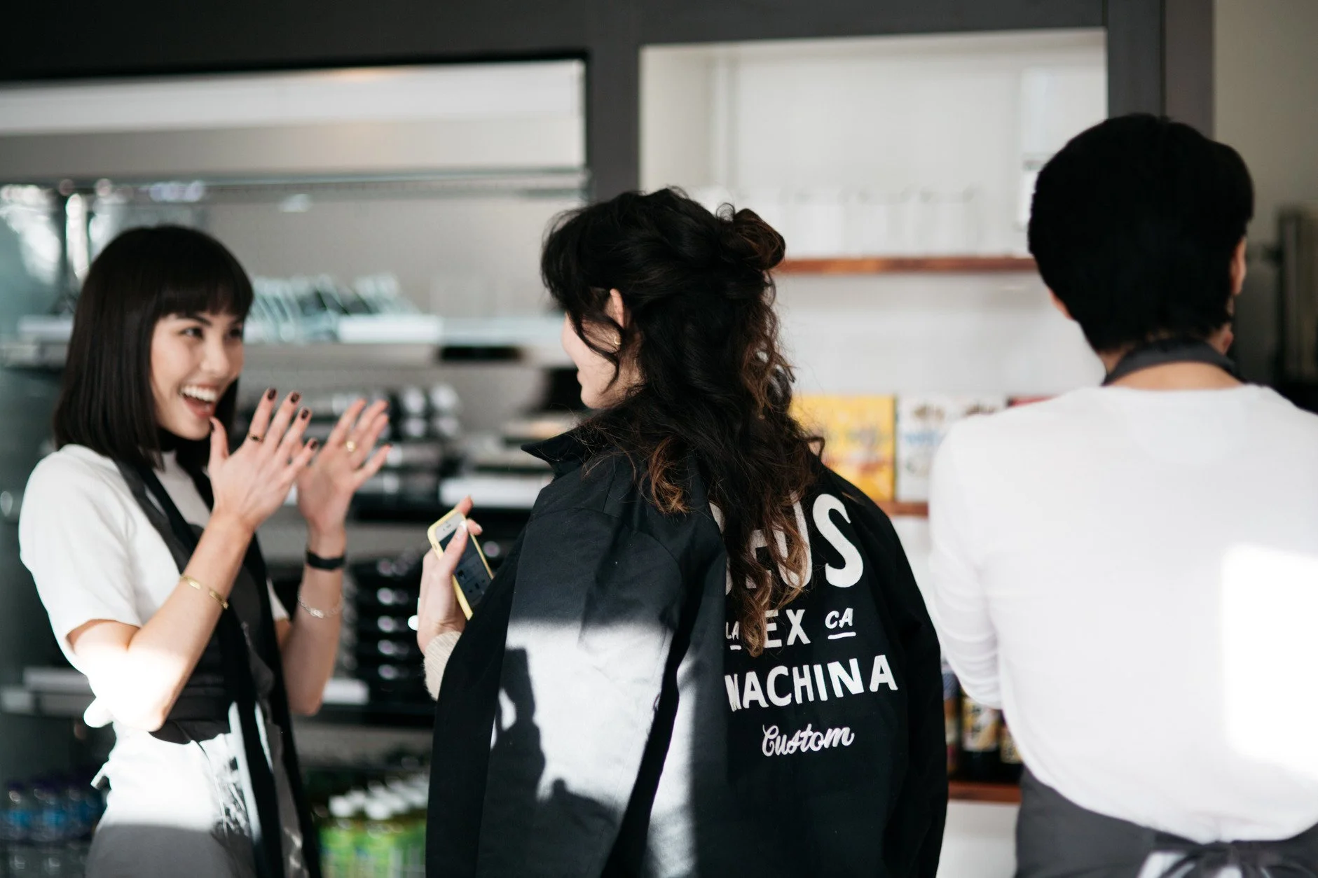 Three people in a retail store engaging in conversation: a smiling young woman with short dark hair and a white shirt, a woman with curly hair wearing a black jacket, and a man with dark hair in a white shirt, with shelves of merchandise in the background.