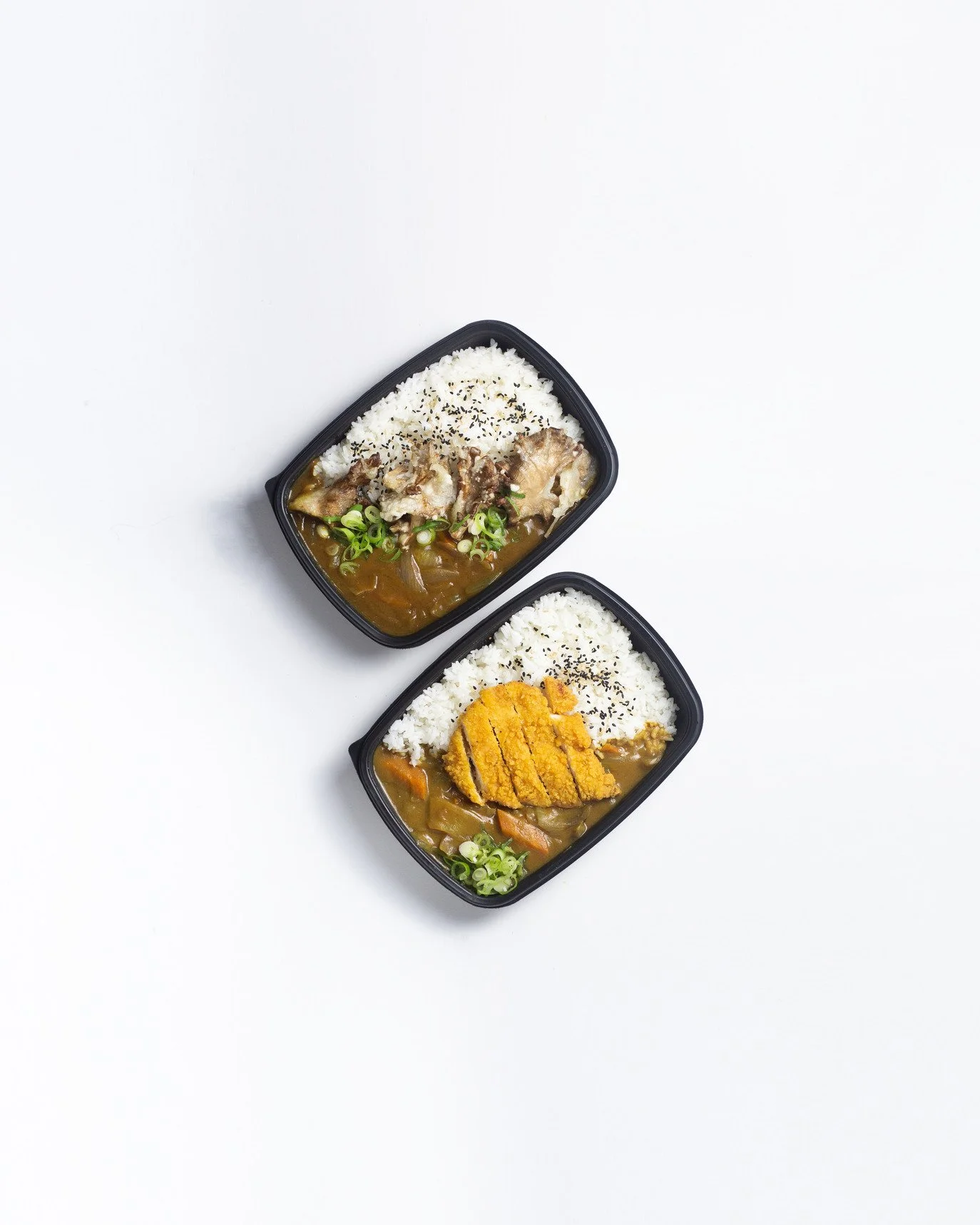 Two bento boxes with Japanese curry, rice, and toppings; one with fried chicken, the other with roasted meat and chopped green onions.