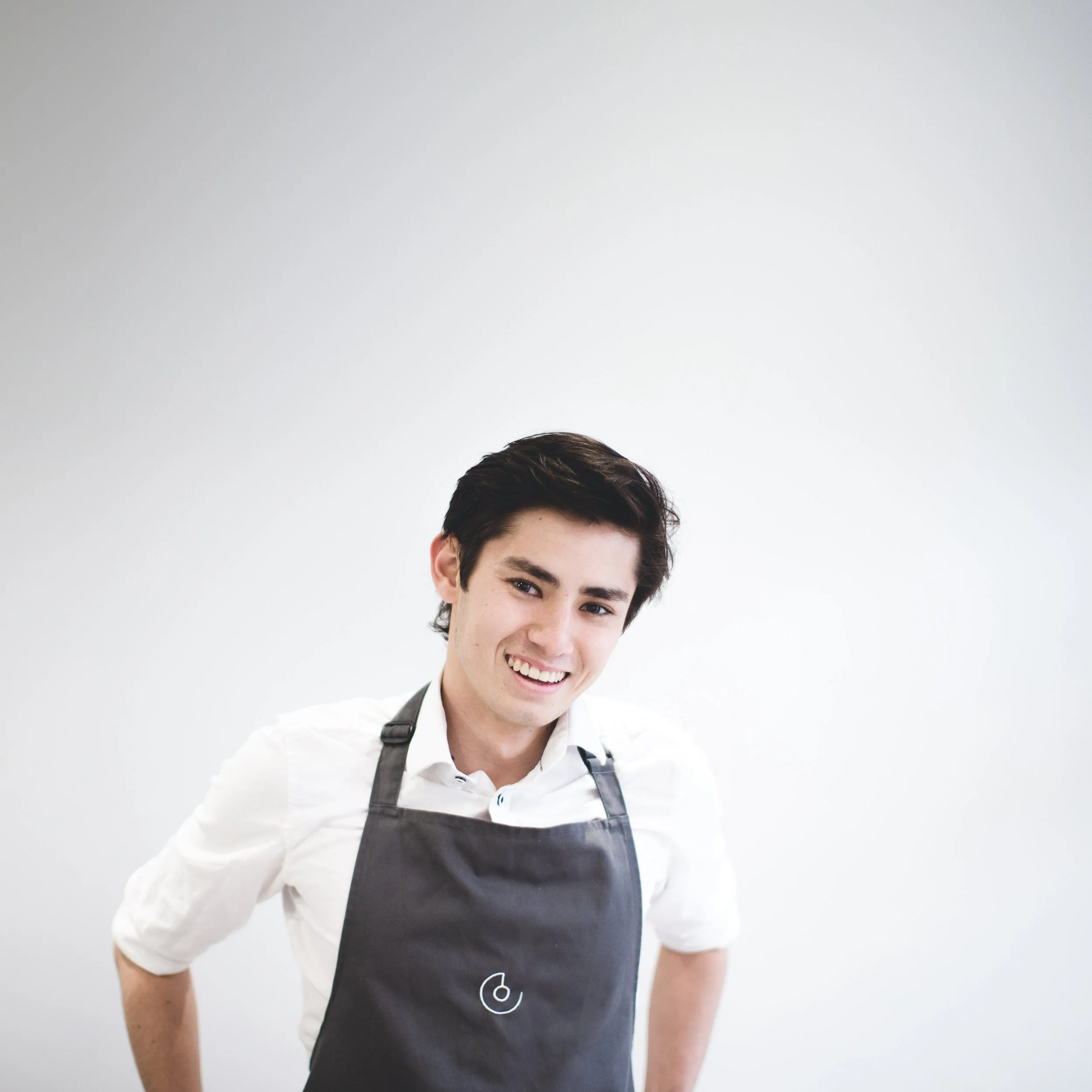 Young man with dark hair smiling, wearing a white shirt and a black apron with a stylized 'G' logo, standing against a plain light gray background.