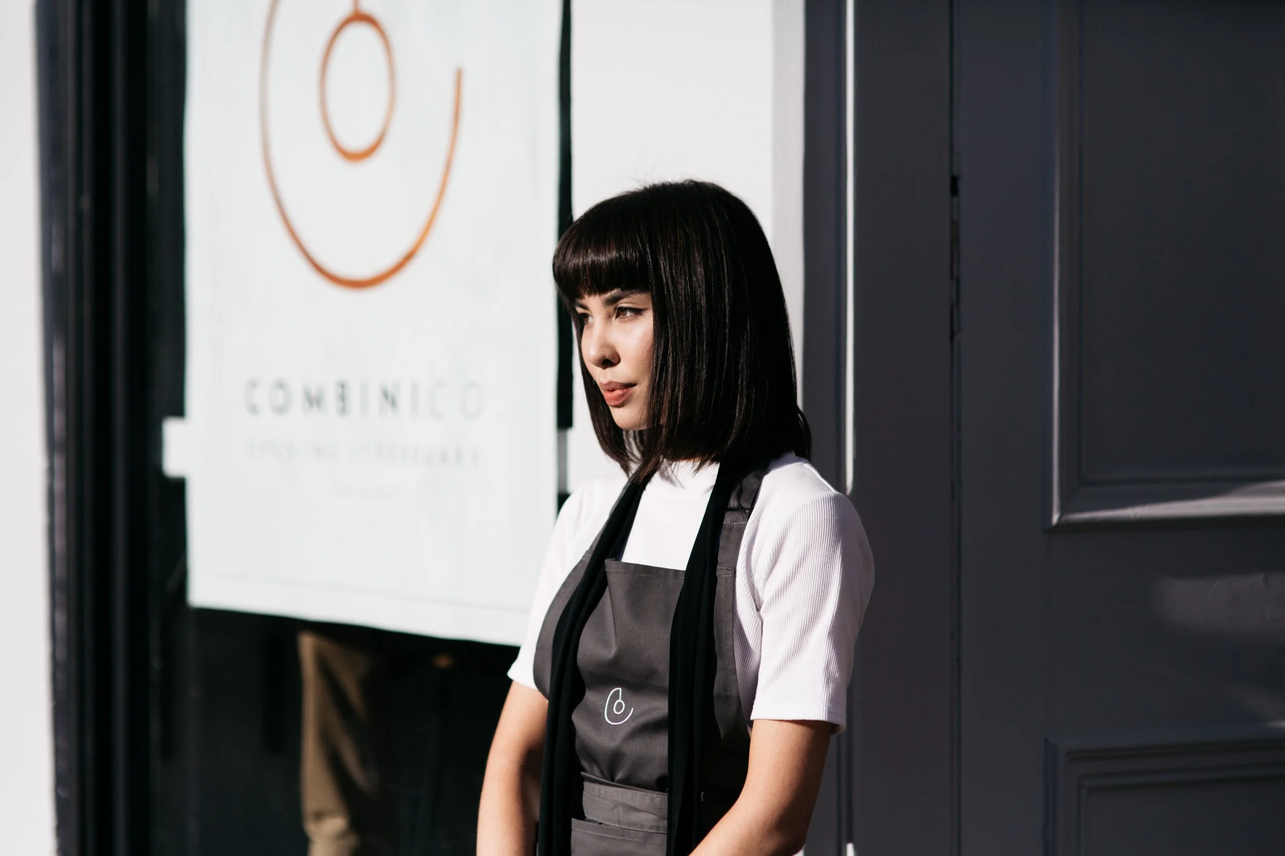 A young woman with black hair and a bob hairstyle standing near a black wall, wearing a white shirt and a dark apron, with a mural or sign on the wall behind her.