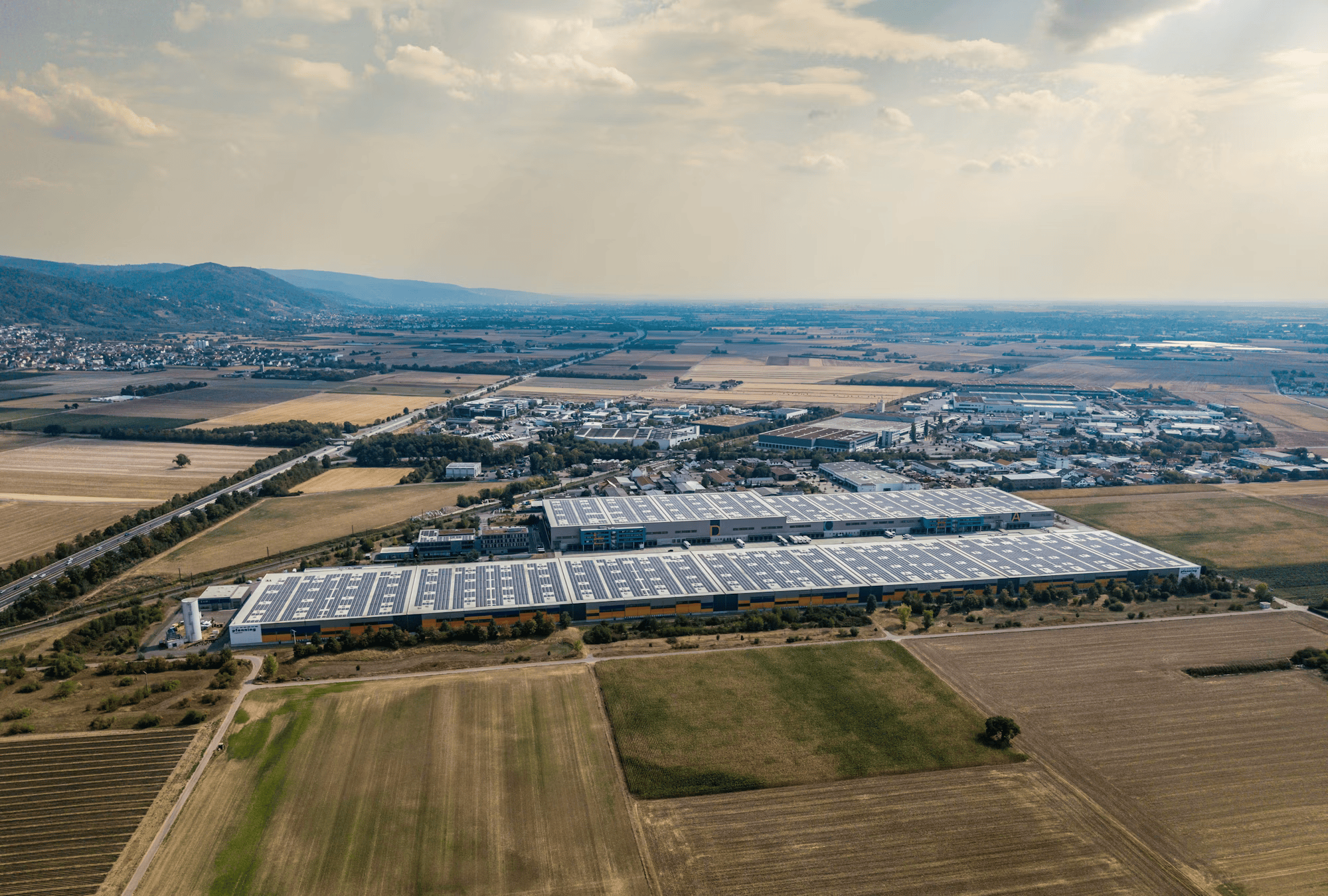 Aerial view of a large industrial complex with solar panels in a rural area, surrounded by farmland and distant mountains under a partly cloudy sky.