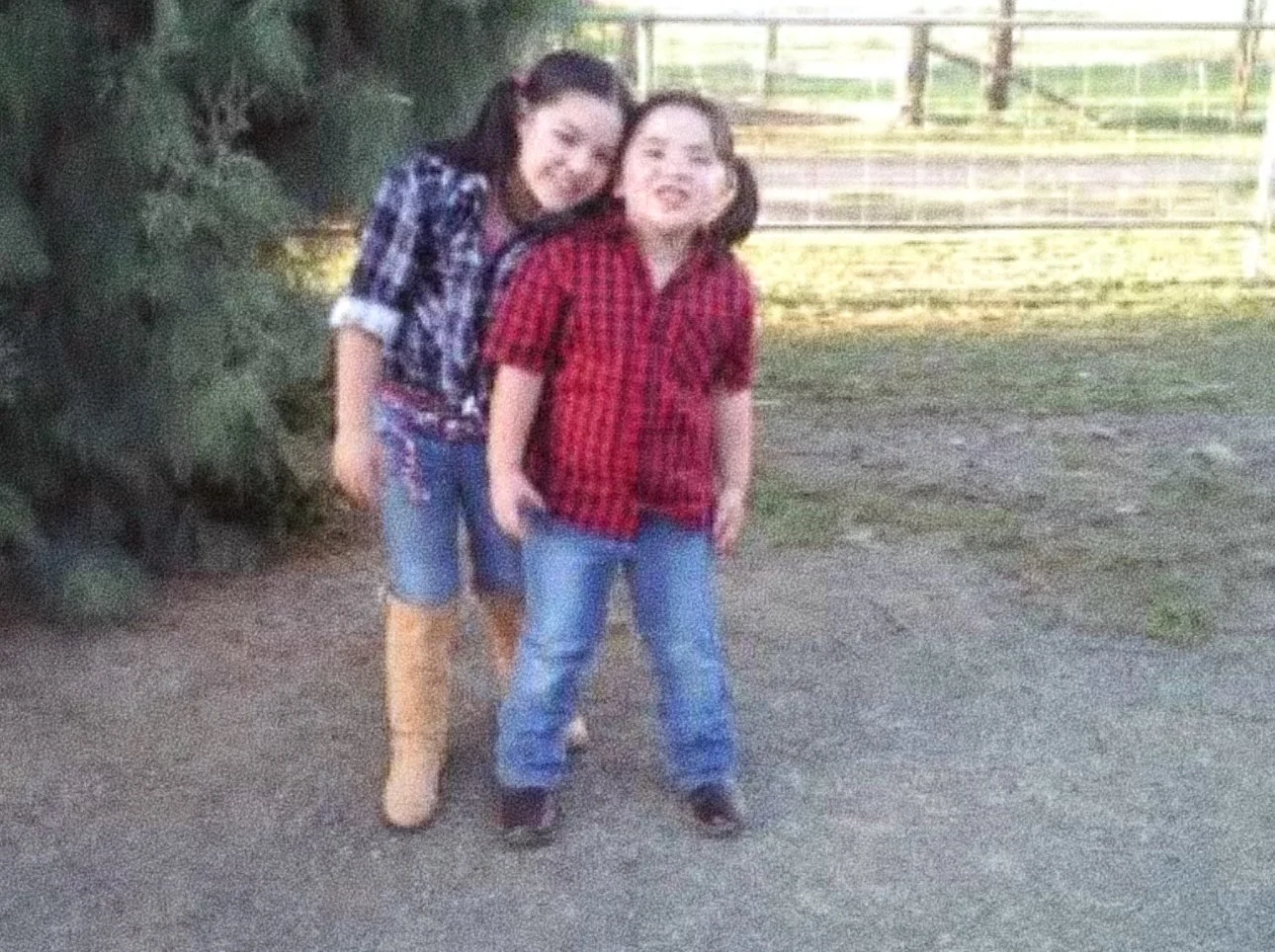 Two children, smiling and standing outdoors on a dirt surface near trees and a fence.