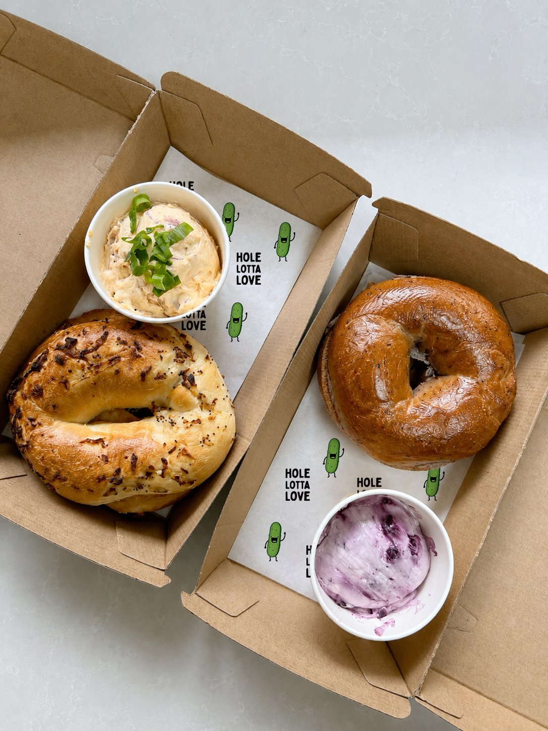 Two cardboard pizza boxes, each containing a bagel and a cup of spread or dip, one with a jalapeño bagel and the other with a plain bagel. The bagels are topped with various ingredients, and the cups contain creamy spreads with one garnished with chopped green onions and the other with swirled purple and white berries.