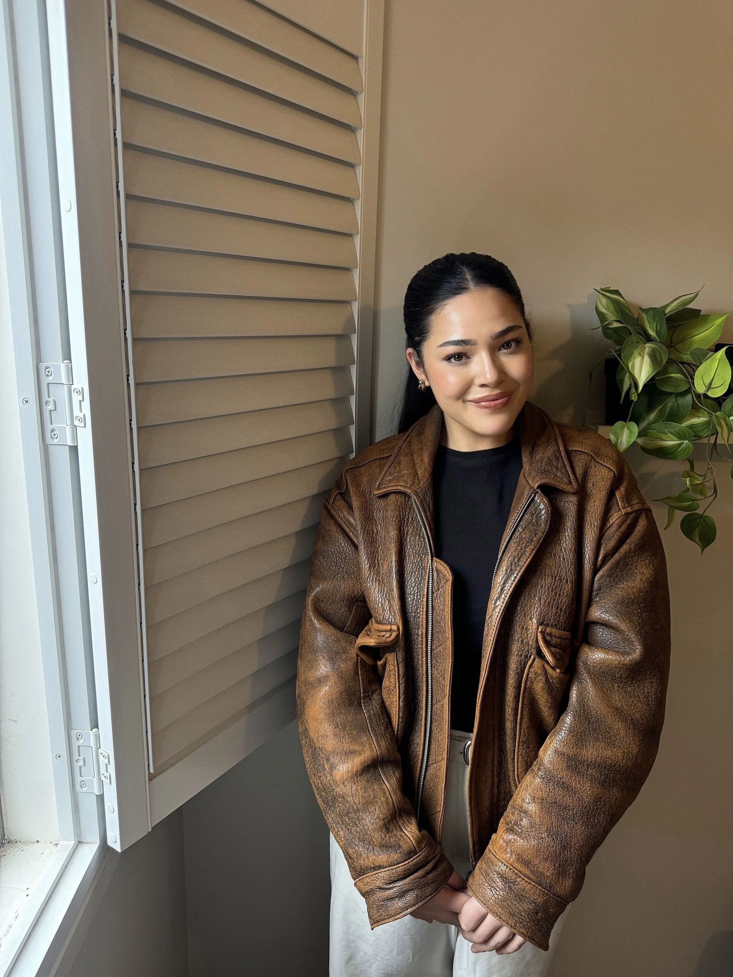 Close-up of a woman wearing a brown leather jacket and black top, standing with her hands clasped in front of her, near a window with closed beige shutters and a houseplant on the right.