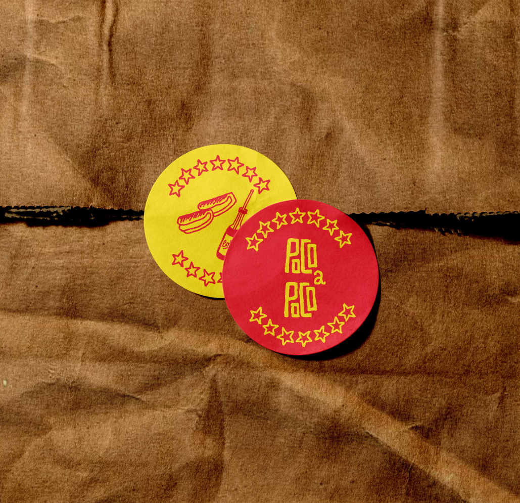 Two circular stickers, one yellow with a hot dog and mustard bottle illustration and the other red with the text 'Polo a Polo' surrounded by star designs, placed on brown paper.