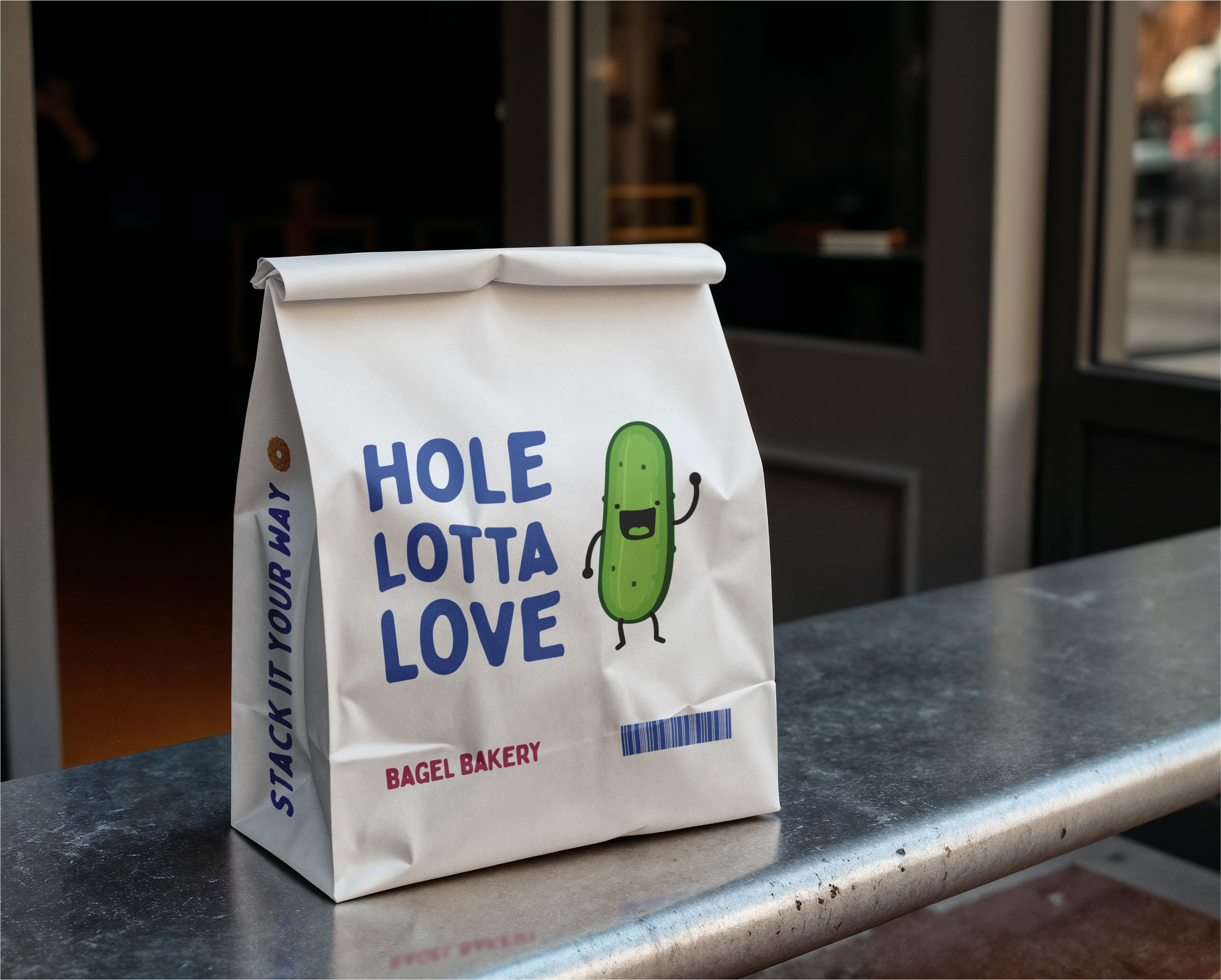 A white paper bag from a bakery with a cartoon cucumber character on it, placed on a stone surface near the entrance of a bakery or cafe.