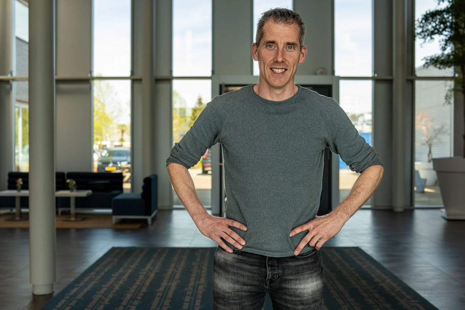 Vrolijke man met hands on hips, binnen in een modern gebouw met grote ramen en buiten zichtbaar groen en auto's.