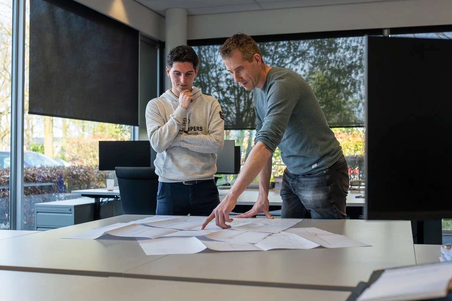 Twee mannen bespreken blauwdrukken op een bureau in een kantoor met grote ramen buiten.