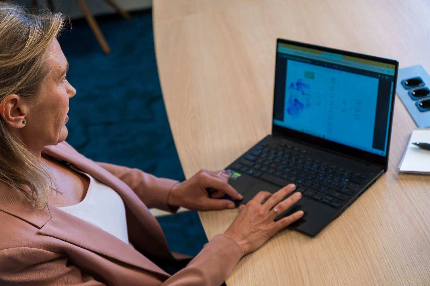 Vrouw werkt aan een laptop op een grote houten tafel, met drie smartphones en een notitieboekje naast haar.
