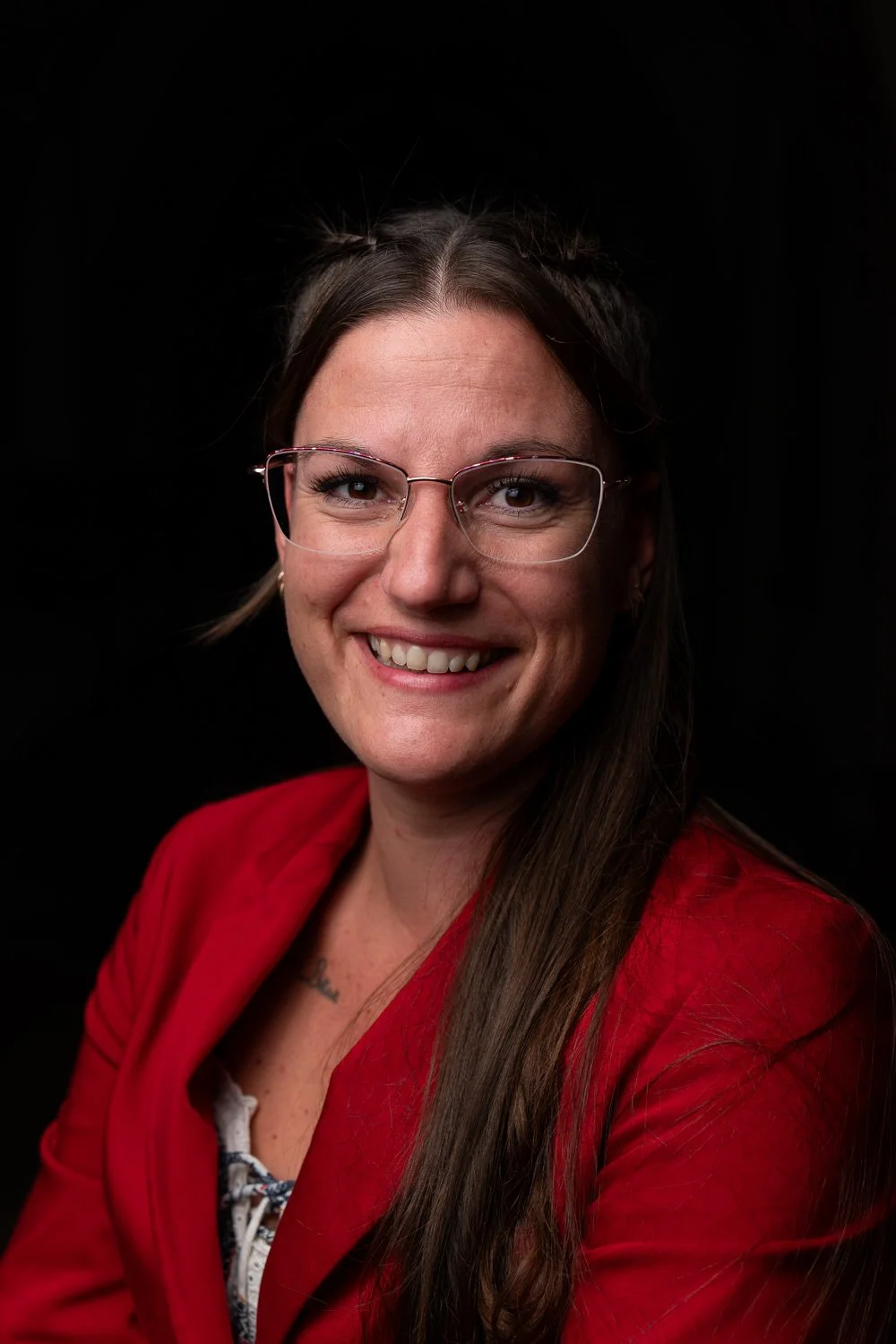 Portret van een lachende vrouw met lang bruin haar, donker bril, rode blazer, tegen een zwarte achtergrond.
