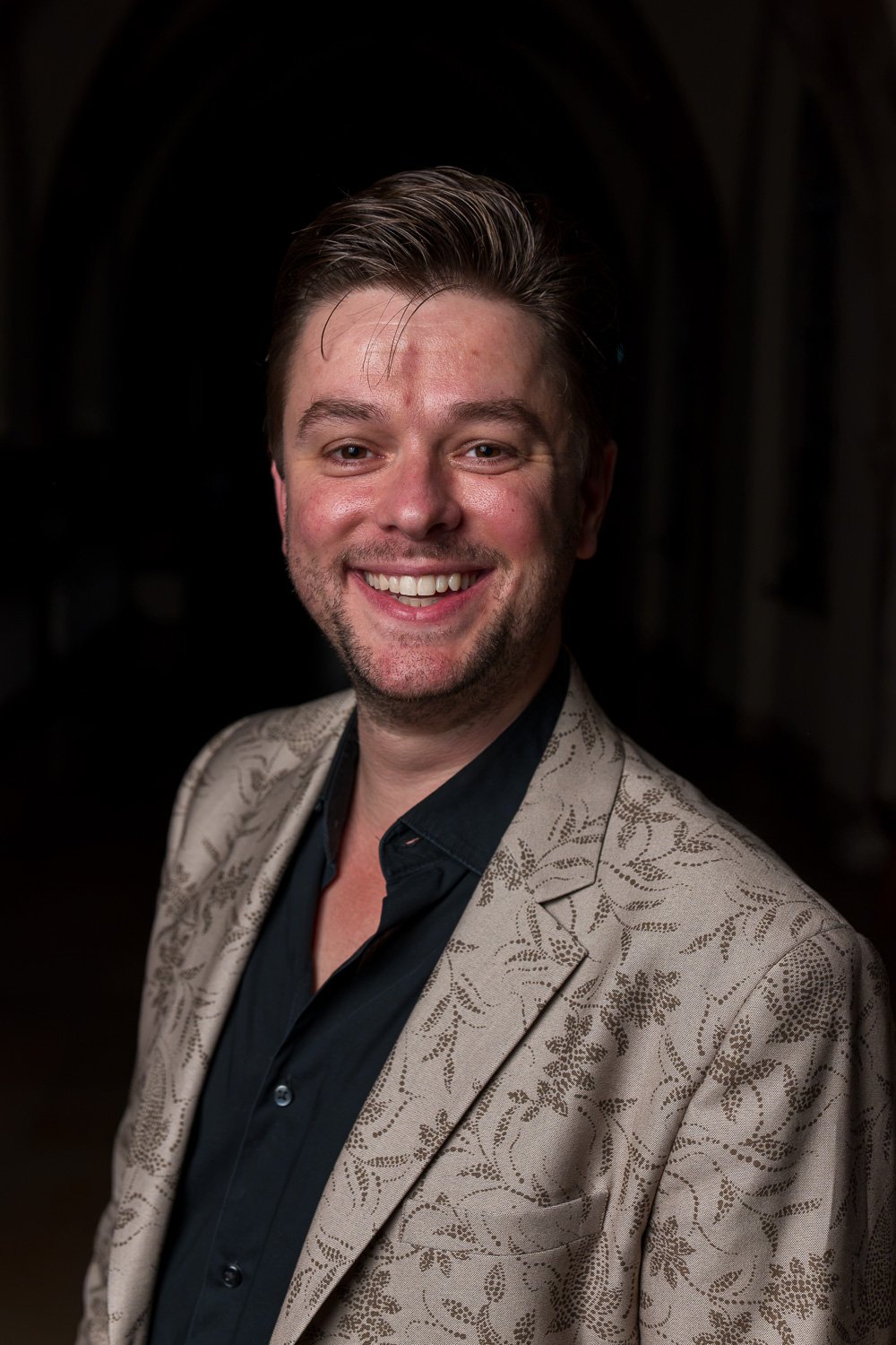 Portret van een lachende man in een beige jas en zwarte shirt, met donker achtergrond.