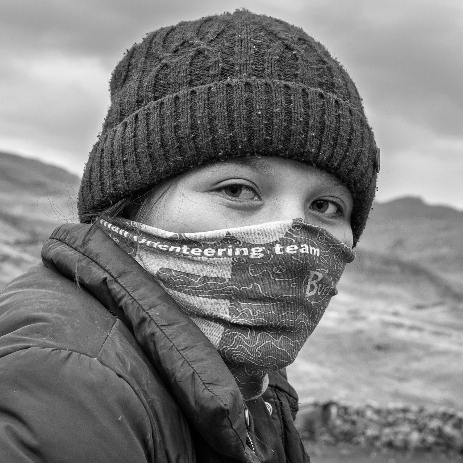 Een persoon met een gebreide muts, een neus- en keelmasker en een een donsjas, met op de achtergrond een berglandschap, in een zwart-witfoto.