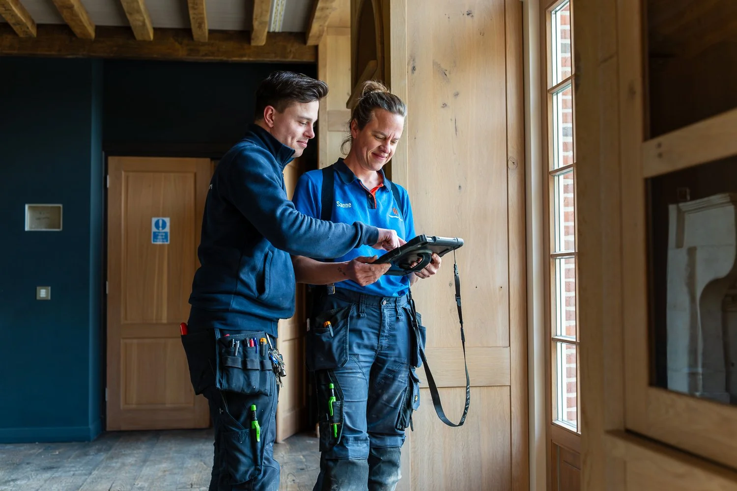Twee monteurs in werkoutfit bekijken een tablet in een houten interieur, mogelijk in een woning of een bouwplaats.