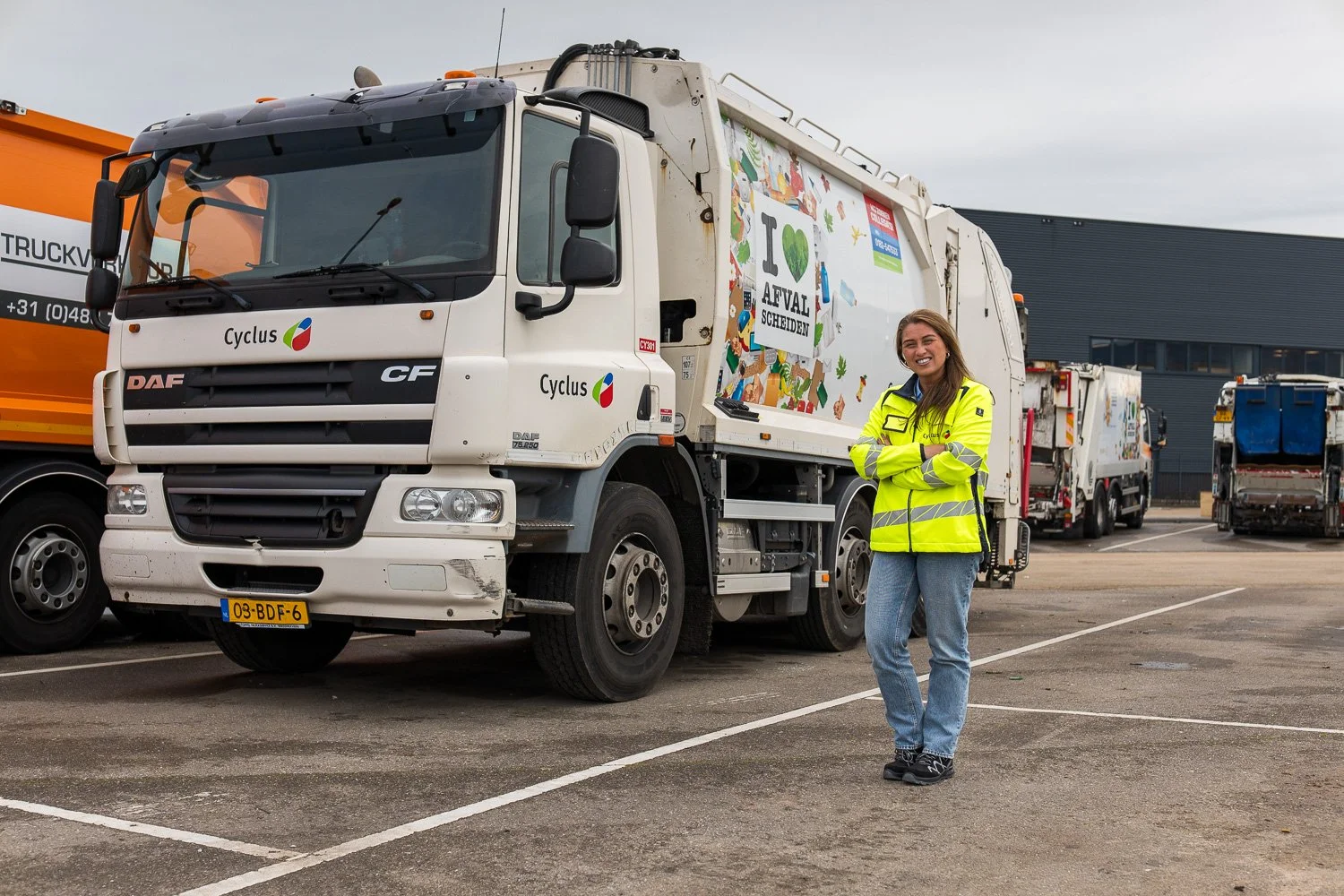 Vrouw in een gele veiligheidsjas die glimlacht en voor een afvalverzamelwagen staat op een parkeerplaats.