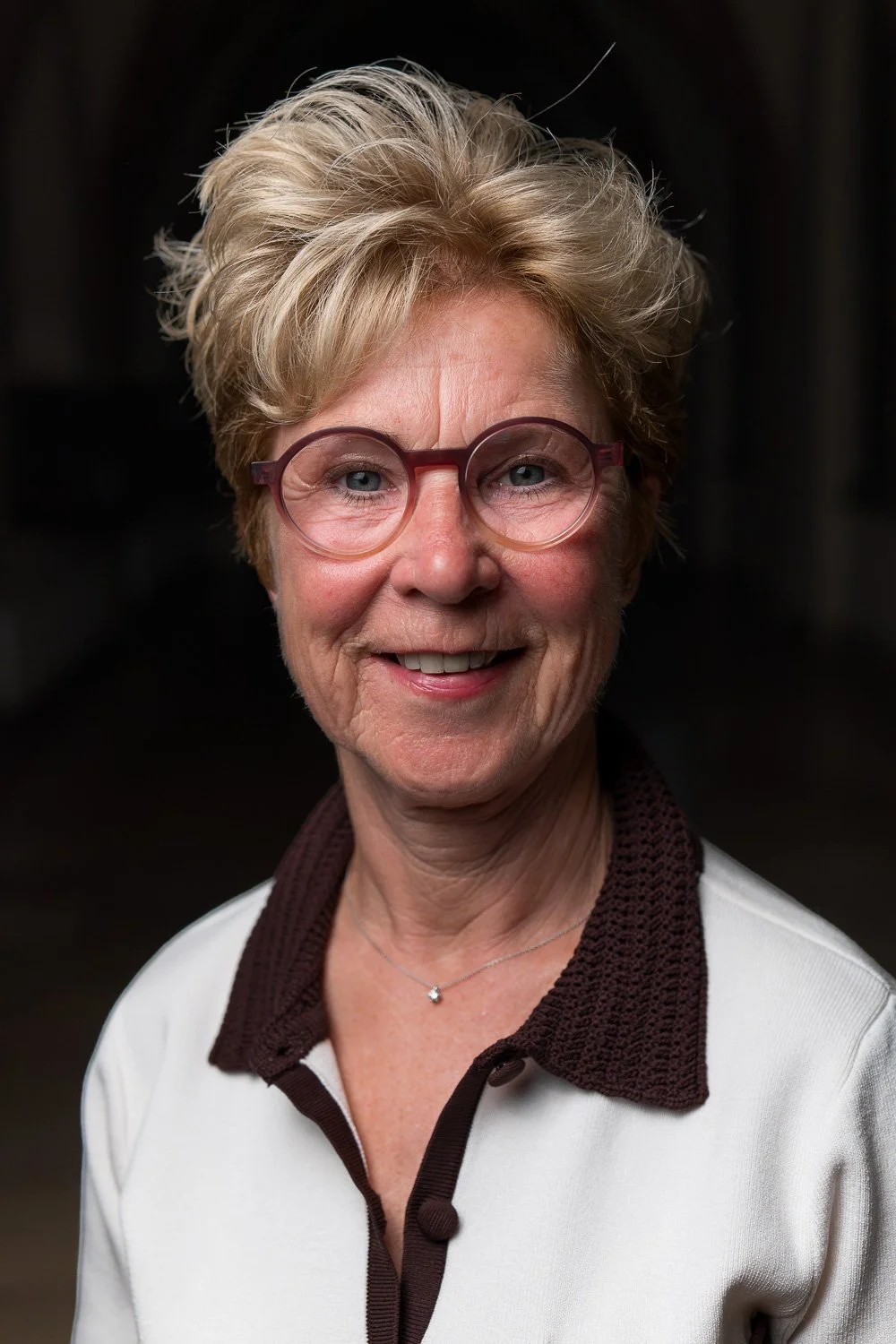 Portret van een lachende oudere vrouw met kort, golvend blond haar, ronde roze brillen, witte blouse met bruin geribbelde col, en een kleine zilveren halsketting.
