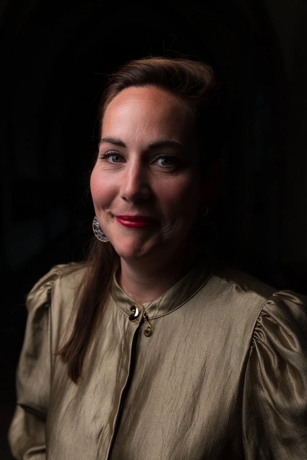Portret van een vrouw met bruin haar, rode lipstick, lichte huid, in een gouden satijnen blouse met pofmouwen, dragen grote gouden oorbellen en een ketting met sluiting, lachend in een donkere achtergrond.