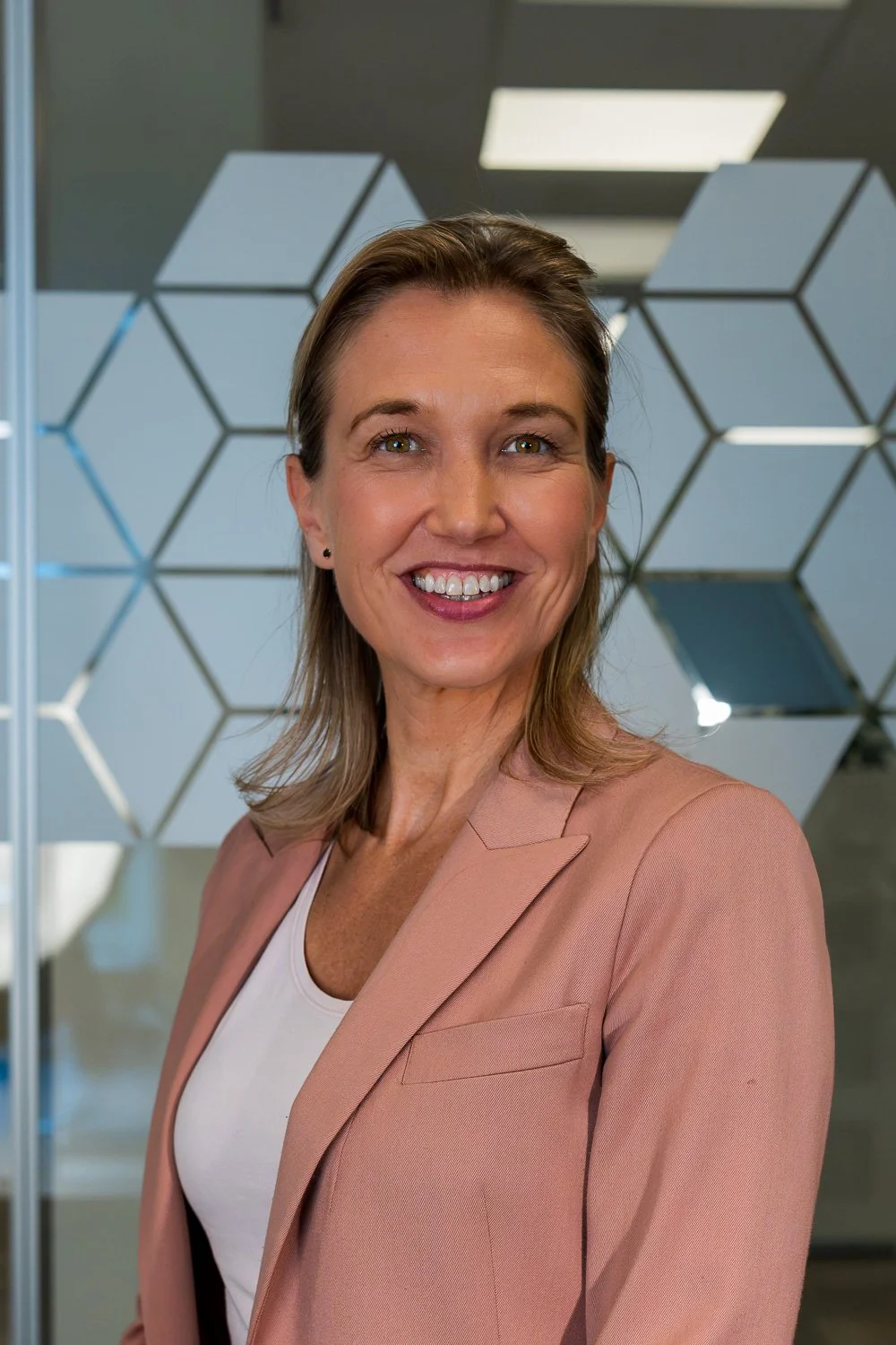 Een glimlachende vrouw in een roze blazer en witte top, staand voor een achtergrond met een geometrisch patroon van hexagonale spiegels.