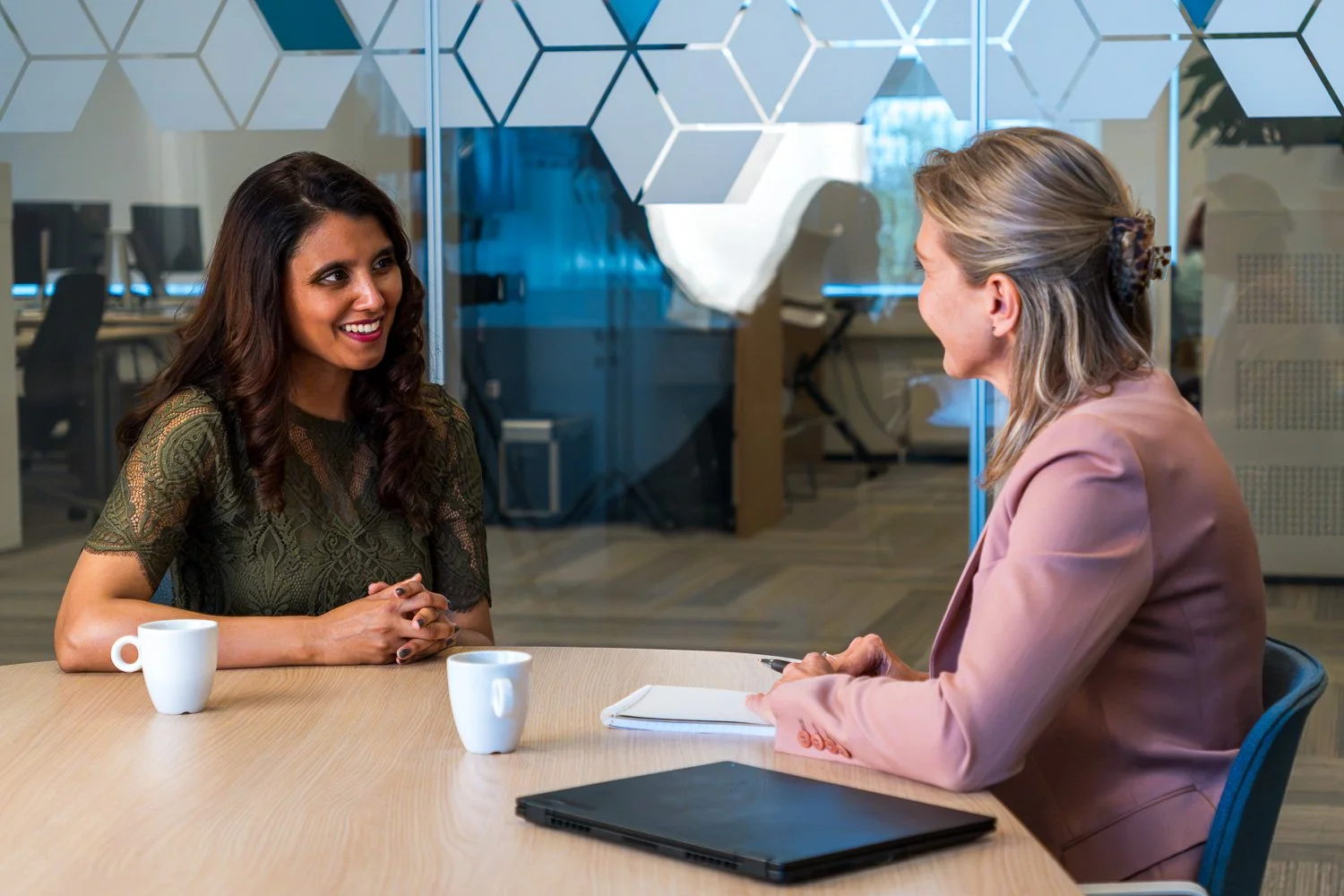 Twee vrouwen hebben een gesprek aan een ronde tafel in een kantoor. Ze glimlachen en er staan twee witte mokken, een open notitieboek en een laptop op tafel.