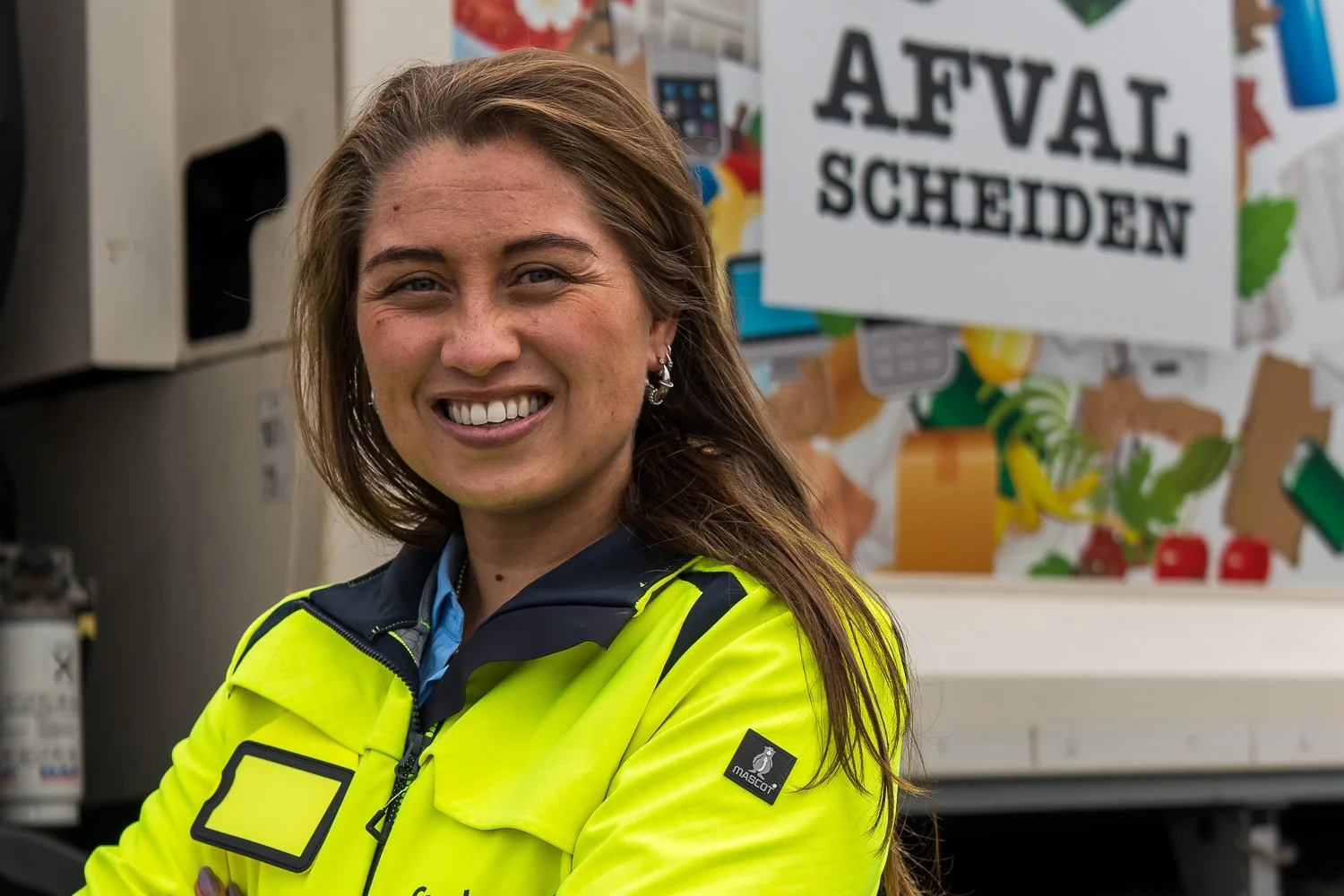 Vrouw in gele jas glimlacht voor een afvalscheidingsstation met een bord dat 'Afval Scheiden' zegt.