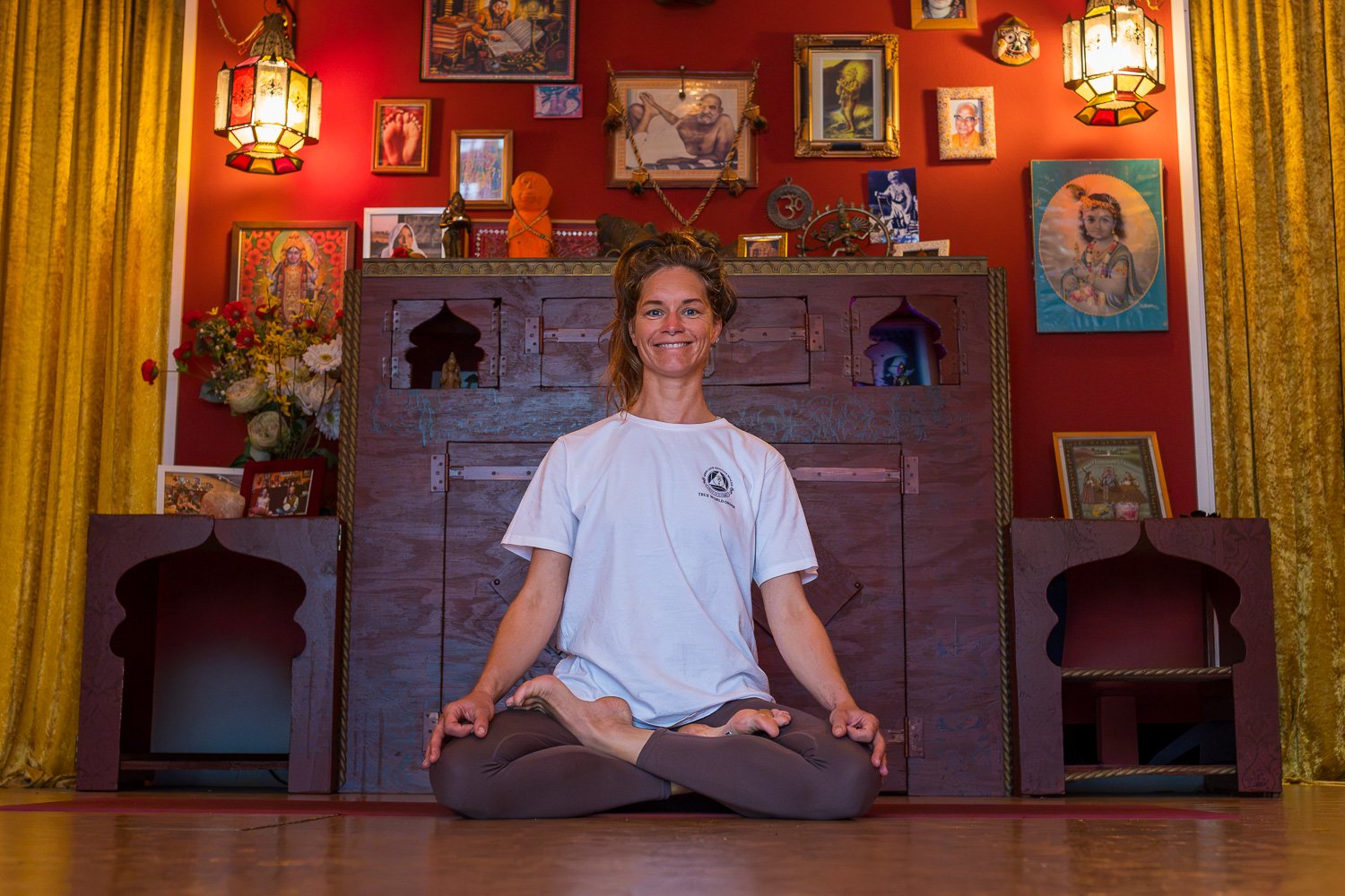 Vrouw in witte yoga kleding zit in lotushouding op de vloer voor een kleurrijk altaar met traditionele Indiase decoraties en afbeeldingen, omringd door gele gordijnen.